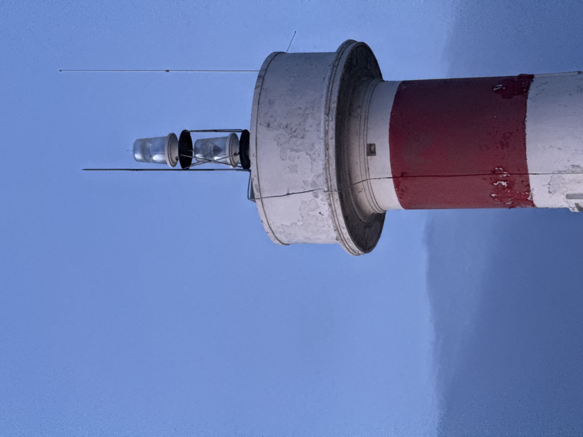 A close-up, low-angle shot captures the top section of a lighthouse against a clear, pale blue sky. The lighthouse is cylindrical, with the visible portion painted in alternating bands of white and red. The white sections are weathered, showing peeling paint, particularly around the top rim. The red band is solid.
At the very top of the lighthouse, there are two clear glass lamps, stacked vertically and encased in a metal frame. Thin metal rods or antennas extend upwards from the lighthouse's structure, one tall and thin on the right, and a shorter, thicker one on the left, just behind the lamps. Faint, dark clouds are visible on the left side of the frame, low on the horizon, contrasting with the bright sky. The overall impression is of a coastal structure, likely at dusk or dawn due to the soft lighting and the pale sky. The image does not depict any people or specific identifiable landmarks of Lý Sơn Special Administrative Region, Vietnam.