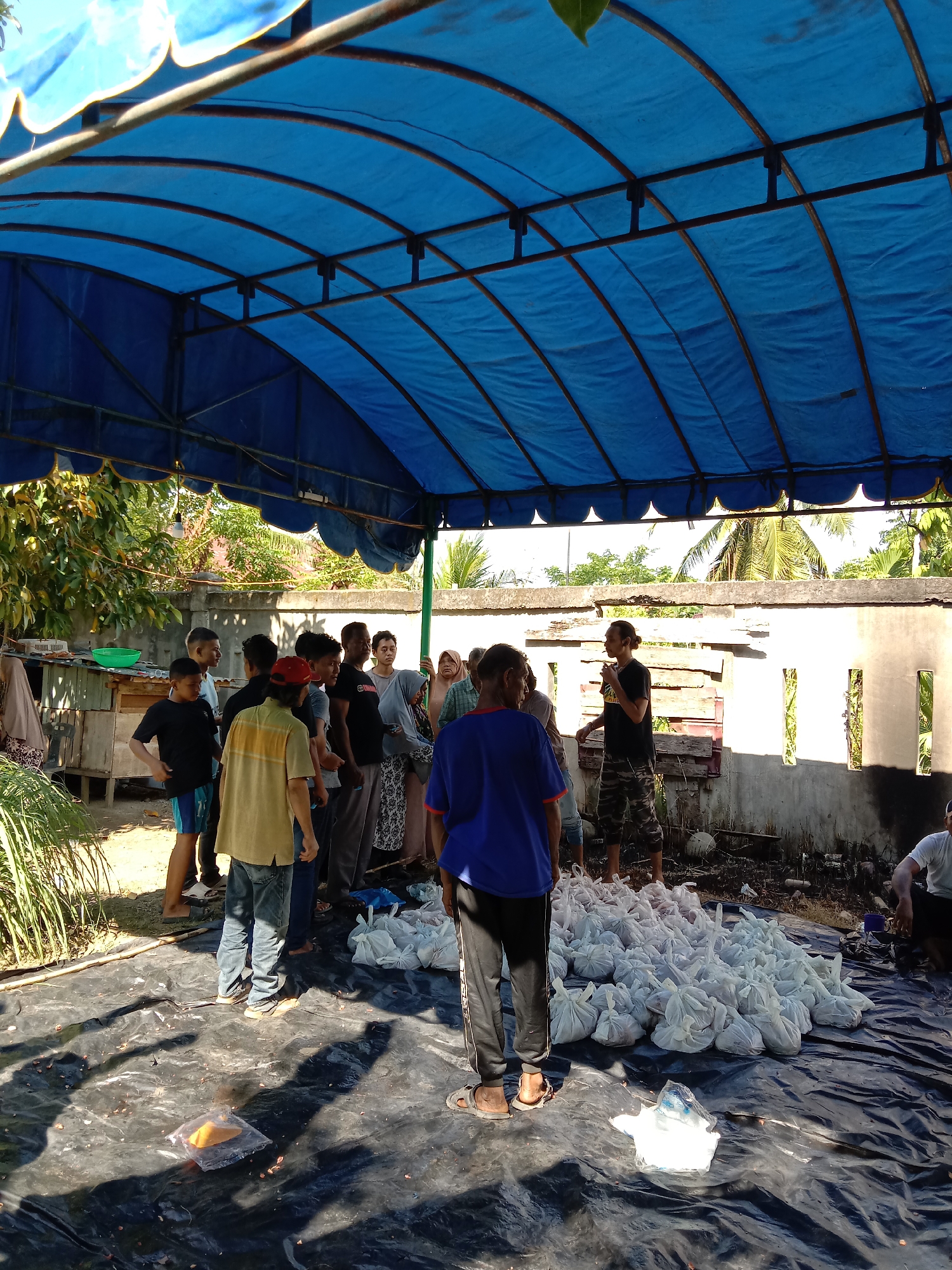 Location: Lhokseumawe, Indonesia.
Scene: A group of people standing under a blue tarp shelter. A large pile of white bags sits on a black tarp. The people are dressed in casual clothing and appear to be engaged in a gathering or distribution of items. A building or wall forms the backdrop, with trees and foliage visible in the background. The environment suggests a community setting.