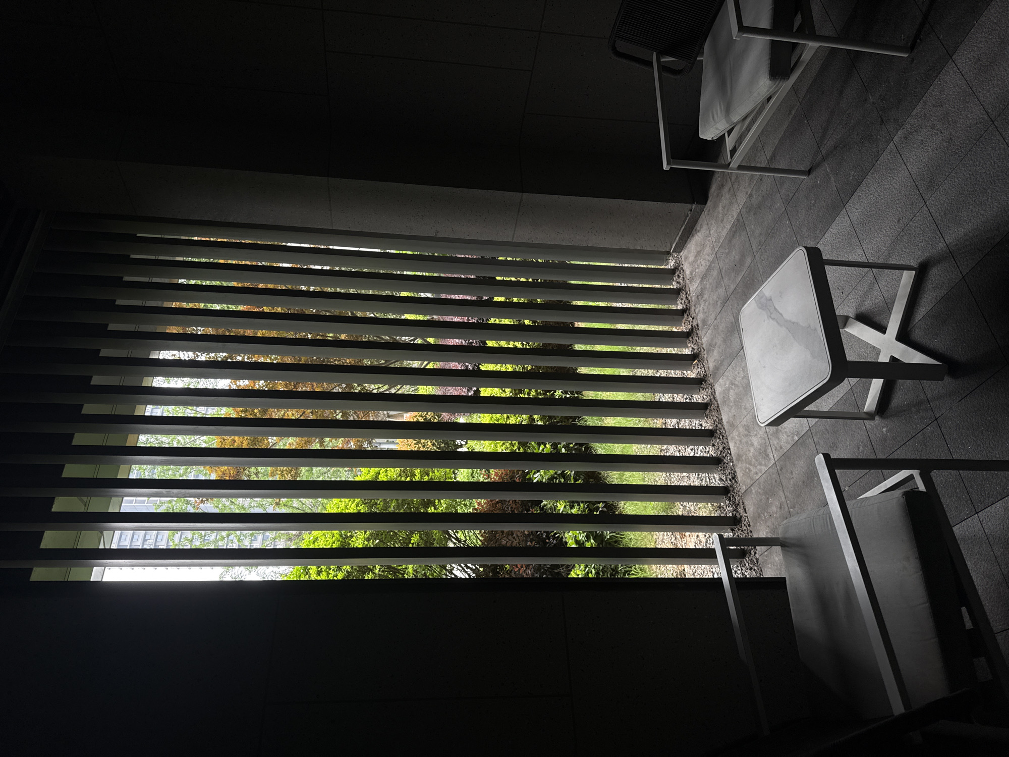 A high-angle photograph captures an unoccupied, modern outdoor seating area in Yiwu, China, bathed in a strong contrast of deep shadows and bright sunlight. The scene suggests a clear, sunny day, likely midday or early afternoon.
On the left, a series of dark horizontal slats or louvers acts as a screen, providing a partial view of a lush green garden. Sunlight streams intensely through the gaps, illuminating varied foliage, including vibrant green plants and hints of reddish-brown leaves. In the background, beyond the garden and filtered through the slats and trees, sections of modern buildings are visible, indicating an urban or suburban environment.
The right side of the image features a contemporary patio or balcony area. A small square table with a light, marble-patterned top and a simple white metal base stands prominently. Two chairs are present: one partially visible in the top right with a dark grey cushion, and another in the bottom right with a lighter grey cushion, both featuring light-colored metal frames. The floor of this seating area is covered with dark grey square tiles, adding texture to the minimalist setting.
No people or activities are depicted, conveying a sense of tranquility and stillness. The composition emphasizes geometric lines and the play of light and shadow, framing the natural vibrancy of the garden against the cool, modern aesthetic of the furniture and structure. No visible text is present.