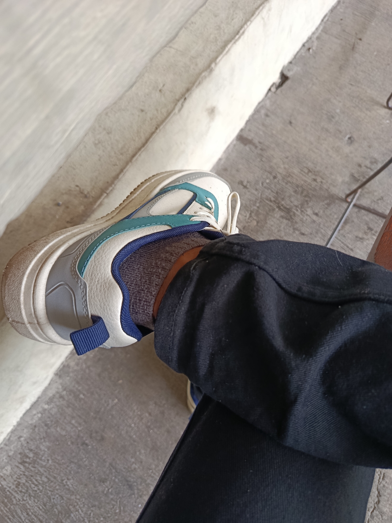 The image shows a close-up view of a person's foot wearing a white, grey, and teal sneaker with a thick sole. The person is also wearing dark blue jeans and grey socks with a blue band. The foot is resting on a concrete surface. The background consists of a textured wall and more concrete. The scene appears to be outdoors, possibly in a public area. There is no specific indication of the city of Cianjur, Indonesia within the image itself.