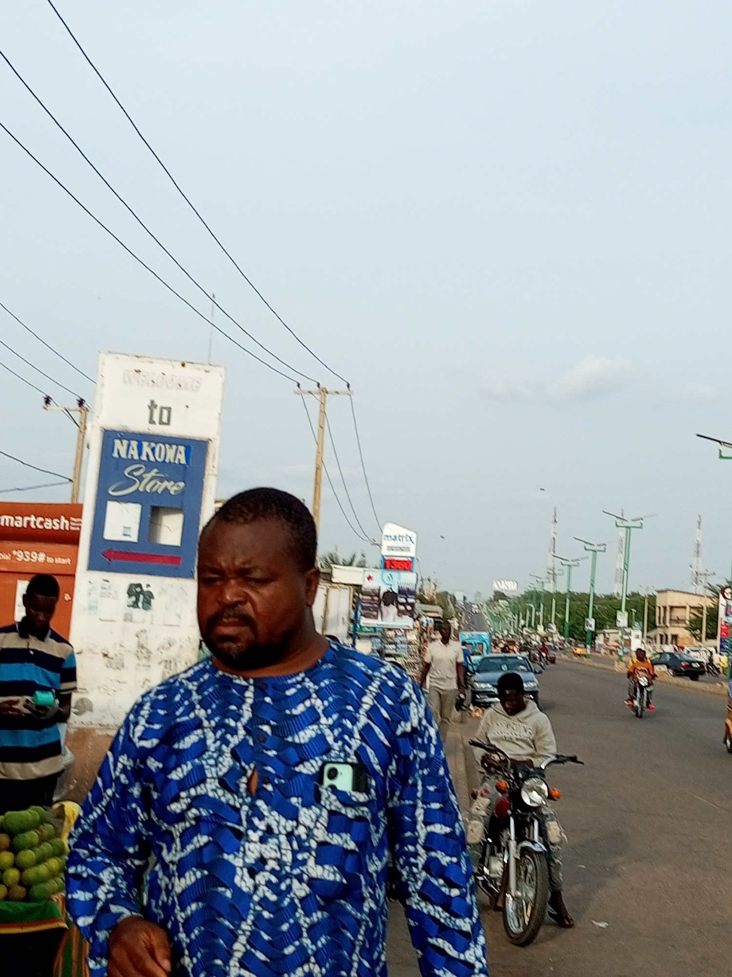 The image depicts a street scene in a city, likely in a tropical or subtropical region, as suggested by the vegetation and clothing styles. The sky is overcast with a few wispy clouds. In the foreground, a man wearing a vibrant blue and white patterned shirt is the central subject. He has dark skin, a beard, and is looking towards the right of the frame. To his left, another man wearing a striped blue and white shirt is standing, holding a green object in his hand. Behind them, a sign reads "WELCOME to NA KONA Store". Further down the street, there is a moderate amount of traffic, including cars and motorcycles, indicating a busy urban environment. Several people are visible on foot and on motorcycles, some engaged in commercial activities, such as a stall selling produce on the left side of the frame. The architecture visible includes various buildings and numerous utility poles with wires crisscrossing the sky. The overall impression is that of a bustling marketplace or a busy commercial street.