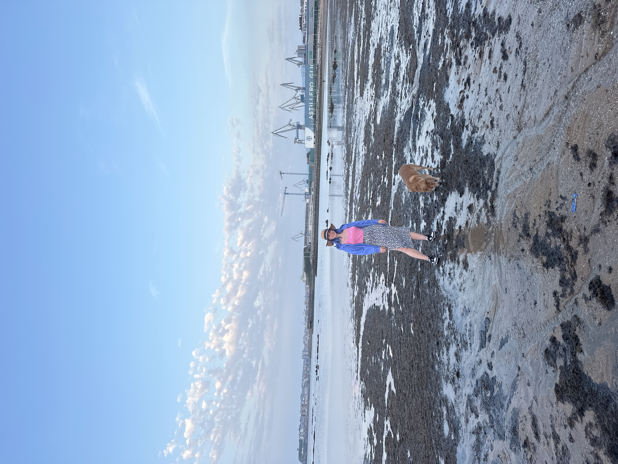The image depicts a scene on a beach, likely at low tide, in Gijón/Xixón, Spain. A woman wearing a straw hat, blue jacket, pink top, and a patterned skirt is standing on the beach. A dog, likely a golden retriever, is nearby. The beach is covered in a mixture of sand and dark mud or algae. In the background, there is a line of industrial cranes and buildings, and the words "ASTILLERO GI" are visible. The sky is blue with some scattered clouds. The composition is vertical, with the beach stretching into the distance.
