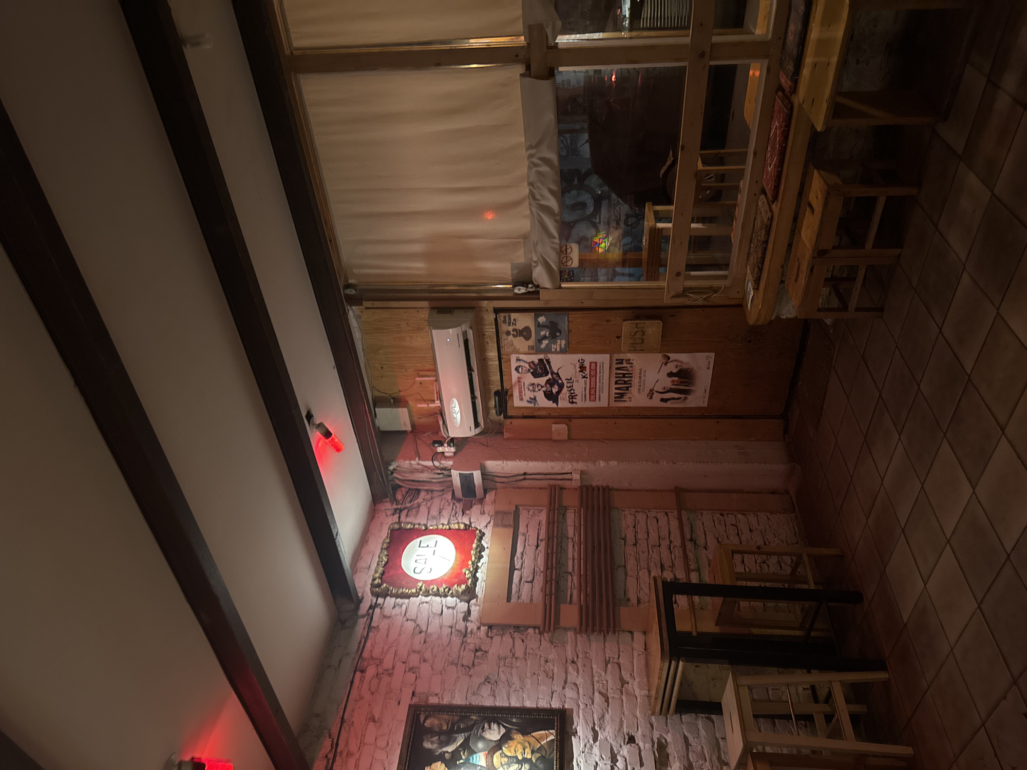 The image shows the interior of a dimly lit establishment. The ceiling has dark wooden beams and red lighting fixtures. The walls are brick, painted white in some areas, and adorned with framed pictures and posters in Bulgarian. An air conditioning unit is visible near the ceiling. The scene includes wooden tables and chairs, some of which appear to be built-in bench seating. Light filters in through a window, showing street graffiti. The location is Sofia, Bulgaria, based on the posters written in Bulgarian.