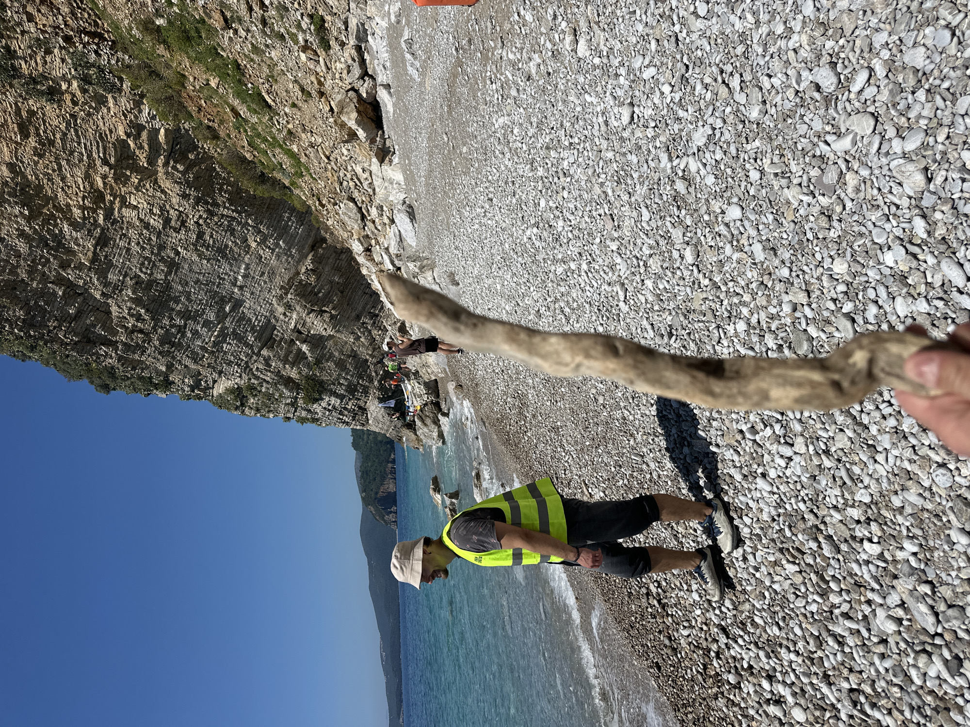 A man in a yellow reflective vest and hat stands on a pebble beach near a cliff and the sea, holding a piece of driftwood. The sea is blue, and the sky is clear. Other people are visible in the background near the cliff. The location is likely near Budva, Montenegro, given the typical landscape.