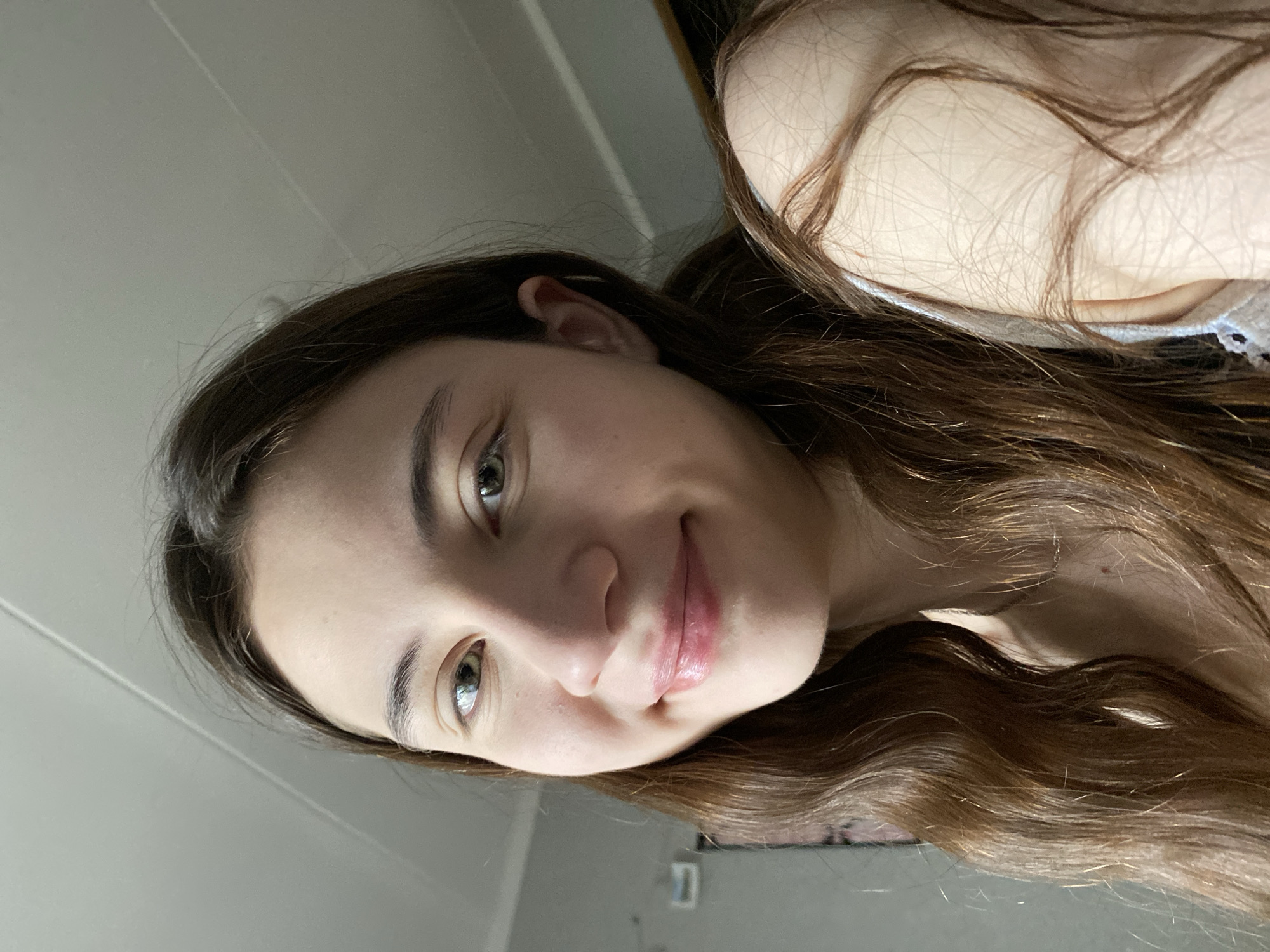 A young woman with long, wavy brown hair and light skin is pictured in a close-up selfie. Her eyes are light-colored. She has a slight smile. She is wearing a light-colored top with lace detailing. The background is a plain wall, possibly in a room. No information suggests the location is Votkinsk, Russia.