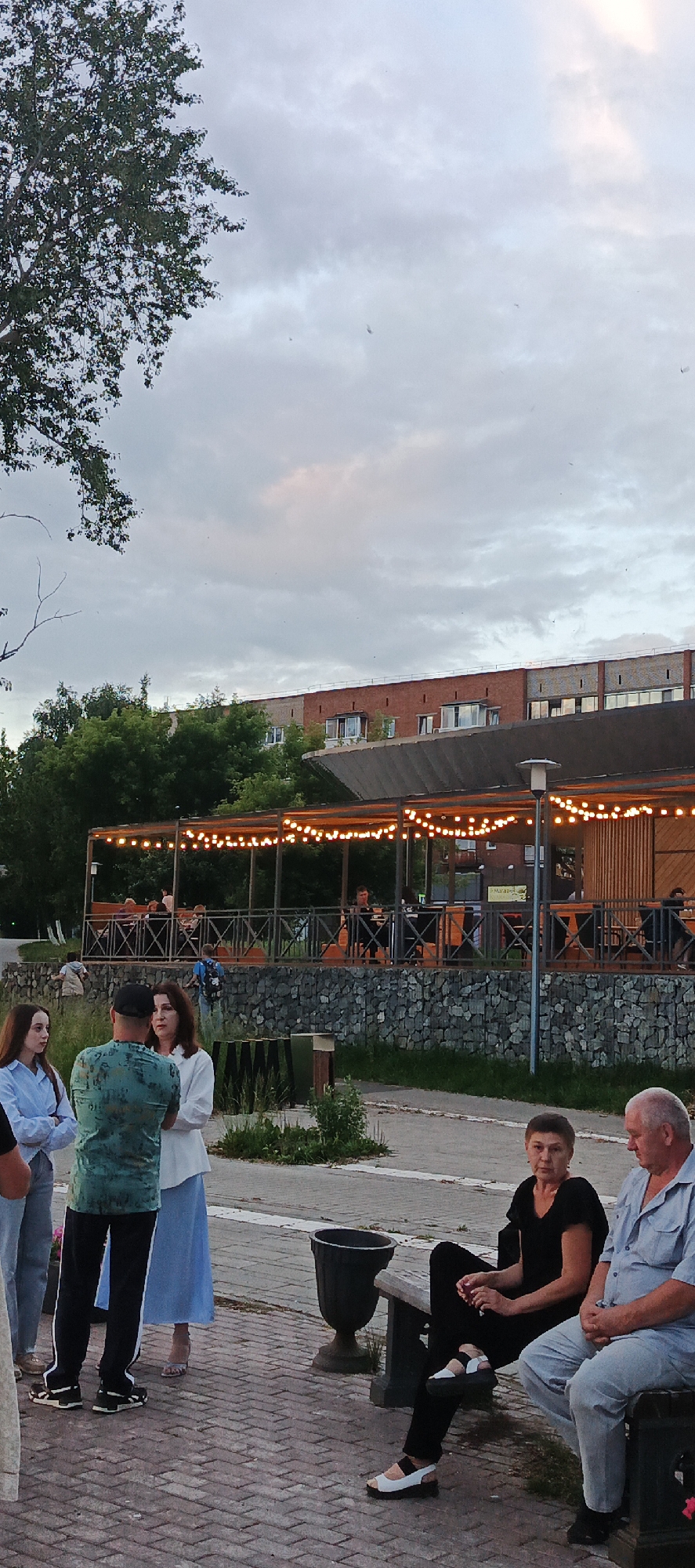 The image captures an outdoor scene, likely an embankment or waterfront area, in Votkinsk, Russia. A group of people are gathered, some standing and conversing, while others are seated on a bench. In the background, a covered outdoor seating area of a restaurant or cafe is visible, illuminated by numerous string lights. Beyond that, a multi-story brick building with windows is present. The environment appears to be a public space with a paved walkway and greenery. The sky is overcast, suggesting it may be evening or a cloudy day.