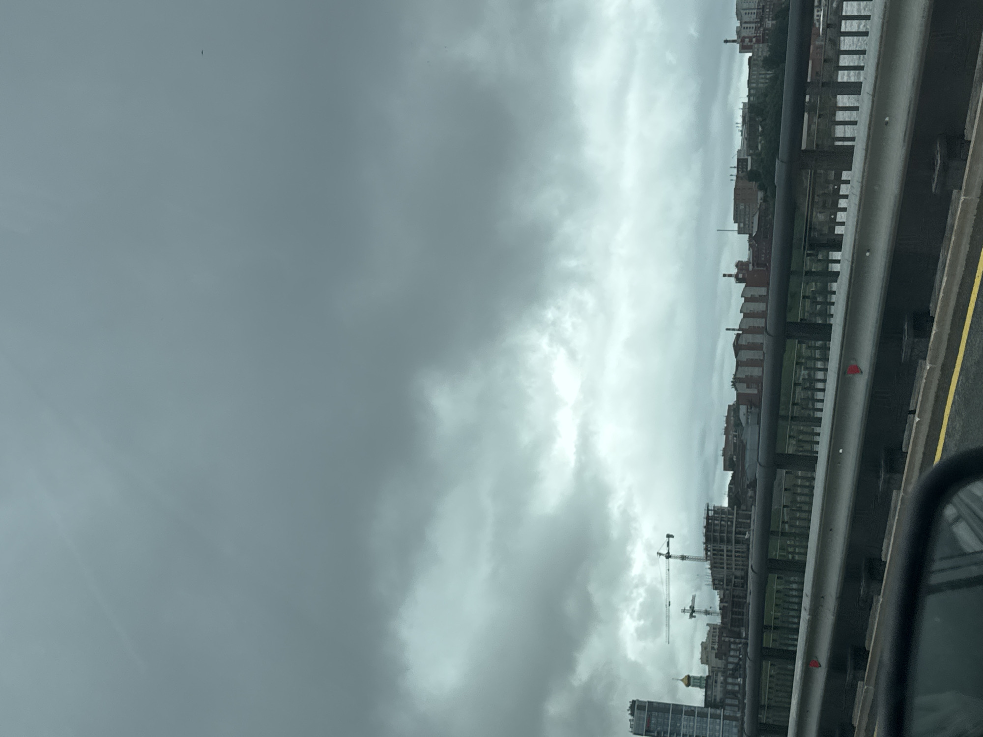 The image, captured from a moving vehicle, shows a view of Perm, Russia under an overcast sky. The foreground includes a highway guardrail and a partial view of a car side mirror. Beyond the guardrail, the cityscape unfolds, marked by buildings of various heights and architectural styles, some of which appear to be residential. Several construction cranes are visible, indicating ongoing development in the area. The background is dominated by a heavy, cloudy sky, obscuring any horizon line. The photograph is oriented vertically. The presence of a waterway is inferred due to the appearance of structures built along what is likely a riverbank. The angle and position suggest this was possibly shot from the Kama Bridge that traverses the Kama River.