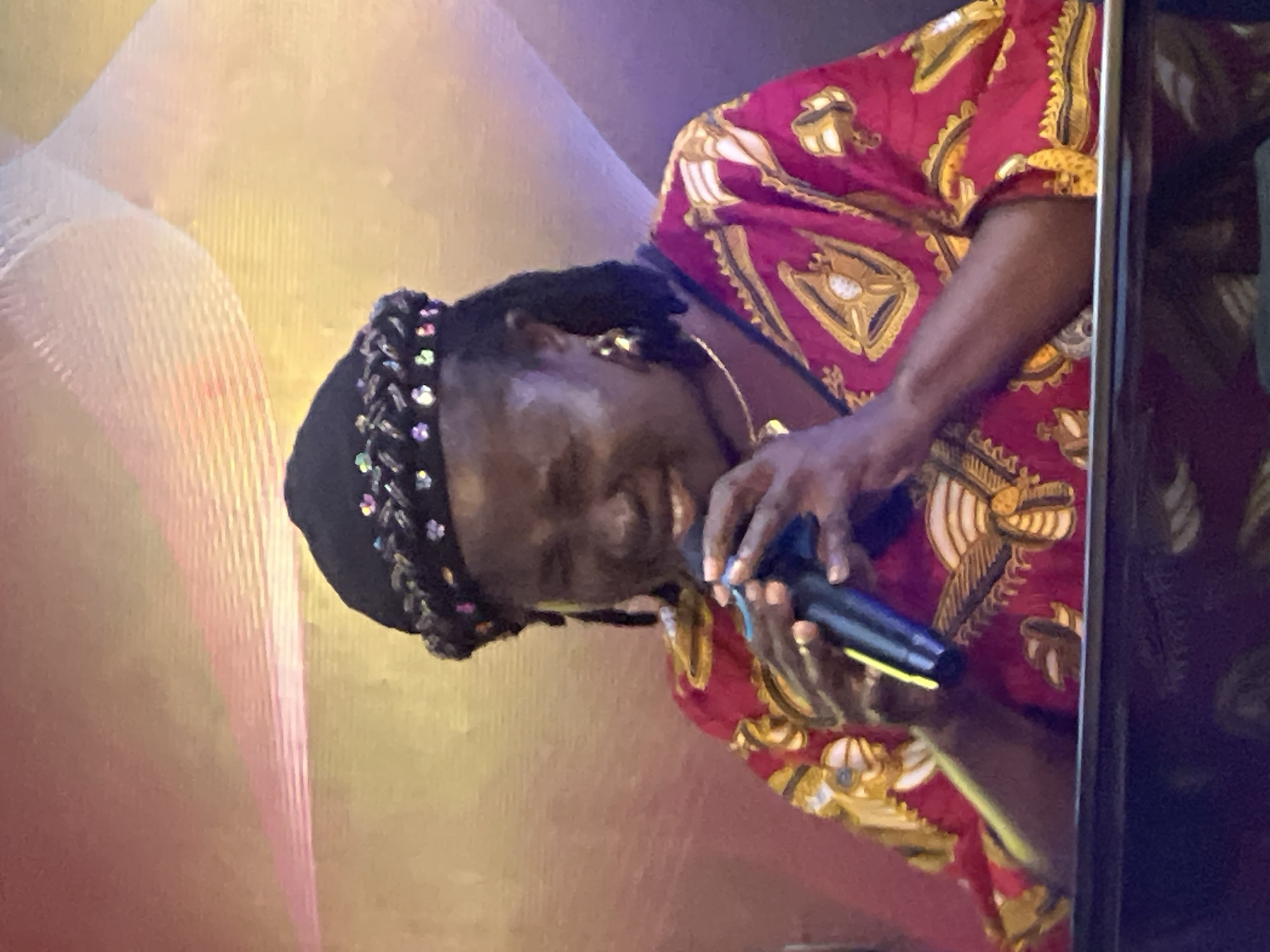 A woman with dark skin and braided hair is singing into a microphone. She is wearing a red and gold patterned garment and a black headwrap adorned with colorful beads. She is wearing large gold hoop earrings and a thin gold necklace. The background is a softly lit, textured wall with abstract wavy lines in shades of yellow and pink. The lighting suggests an indoor setting, possibly a stage or performance venue in Nkpor, Nigeria.