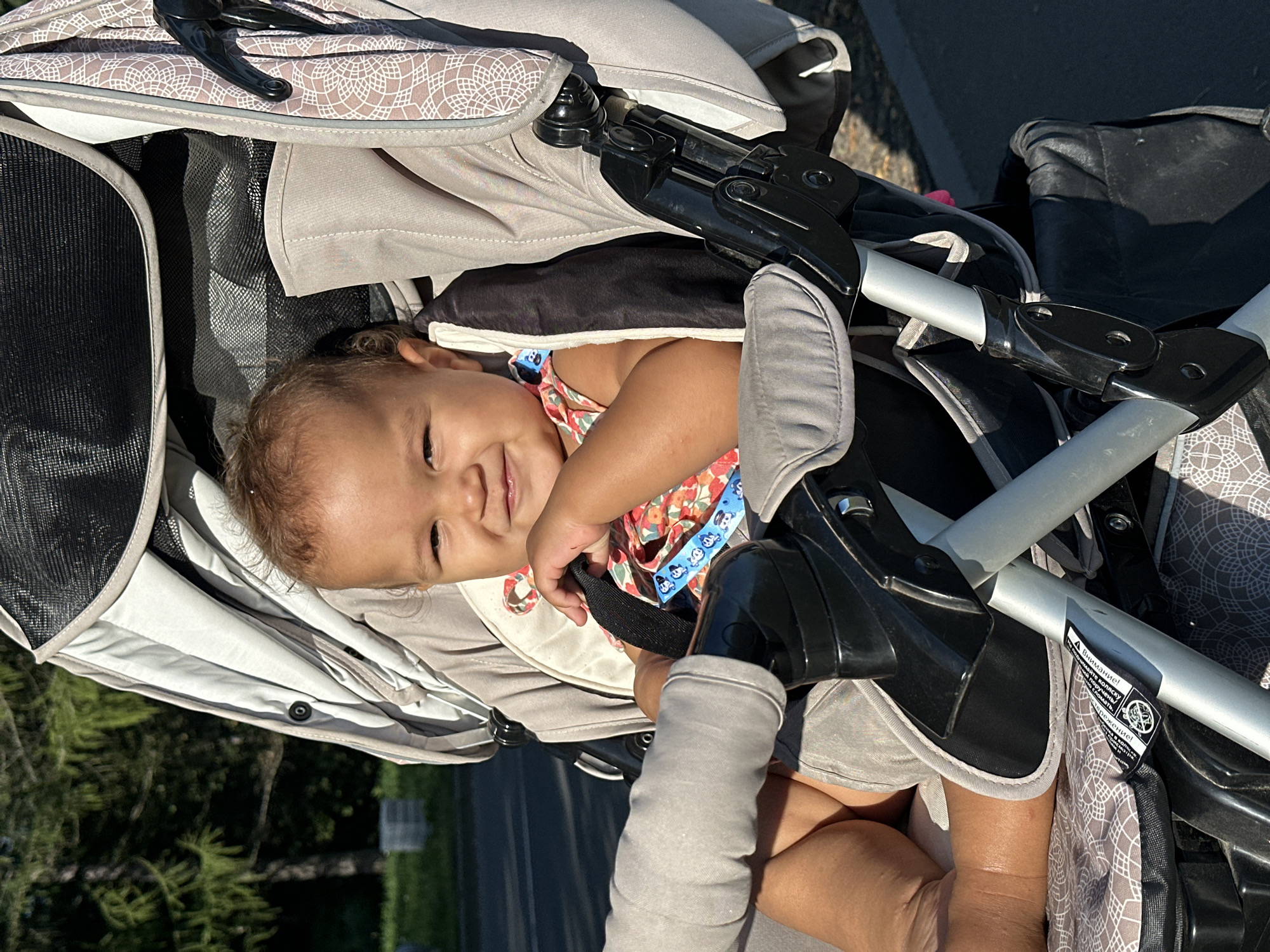 The image shows a close-up of a baby sitting in a stroller outdoors. The baby, appearing to be of mixed ethnicity with light brown hair, is smiling and looking towards the camera. They are wearing a colorful, patterned sleeveless top. The stroller is gray with some patterned fabric accents and black plastic components. The background is blurred, but appears to be a park or outdoor area with green trees and sunlight. A portion of an adult's arm, also appearing to be of mixed ethnicity, is visible on the left side of the frame, resting on the stroller. The lighting suggests it is daytime, likely afternoon. The stroller has safety straps and a sunshade visible. There is a sticker on the stroller frame that appears to have text in Cyrillic. It is not possible to determine the specific location within the city of Petrozavodsk, Russia, from the visual information provided.