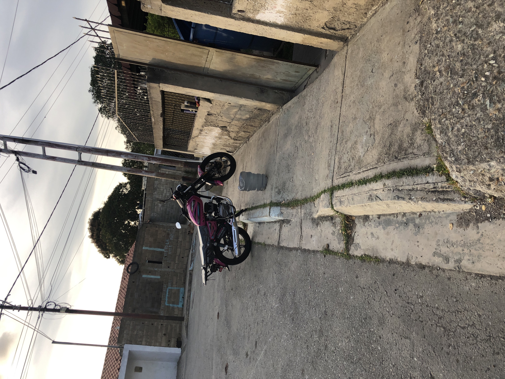 The image depicts a street scene in Altagracia, Venezuela. A pink and black motorcycle is parked on the sidewalk next to a plastic water bottle. A utility pole with numerous wires is visible on the left side of the frame. Buildings are on both sides of the street, constructed of concrete and brick. One building has blue painted squares and graffiti, a sign reading "pistols are not allowed." The street itself is paved and appears to be in a residential area. The sky is overcast.
