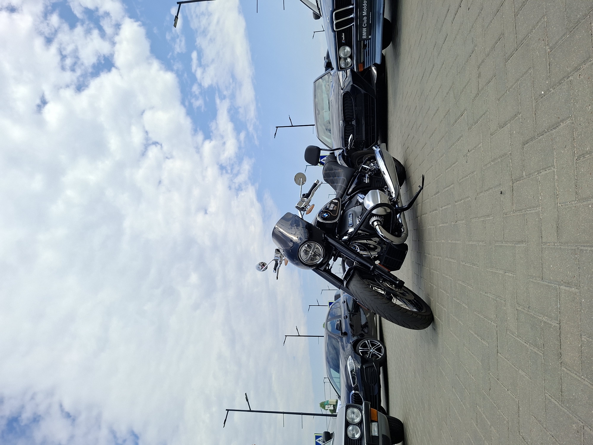 A black BMW R 18 motorcycle is parked in the foreground on a paved surface. The motorcycle is facing left, with its front wheel turned to the left. A windshield, chrome handlebars, and exhaust pipes are visible. In the background, several cars are parked: a gray car on the far left, a black BMW sedan in the middle, and a light blue BMW sedan on the right. The light blue BMW has a license plate that reads "BMW Club Moldova". Streetlights and a cloudy blue sky are visible in the background. The scene appears to be an outdoor parking lot in Stăuceni, Moldova.