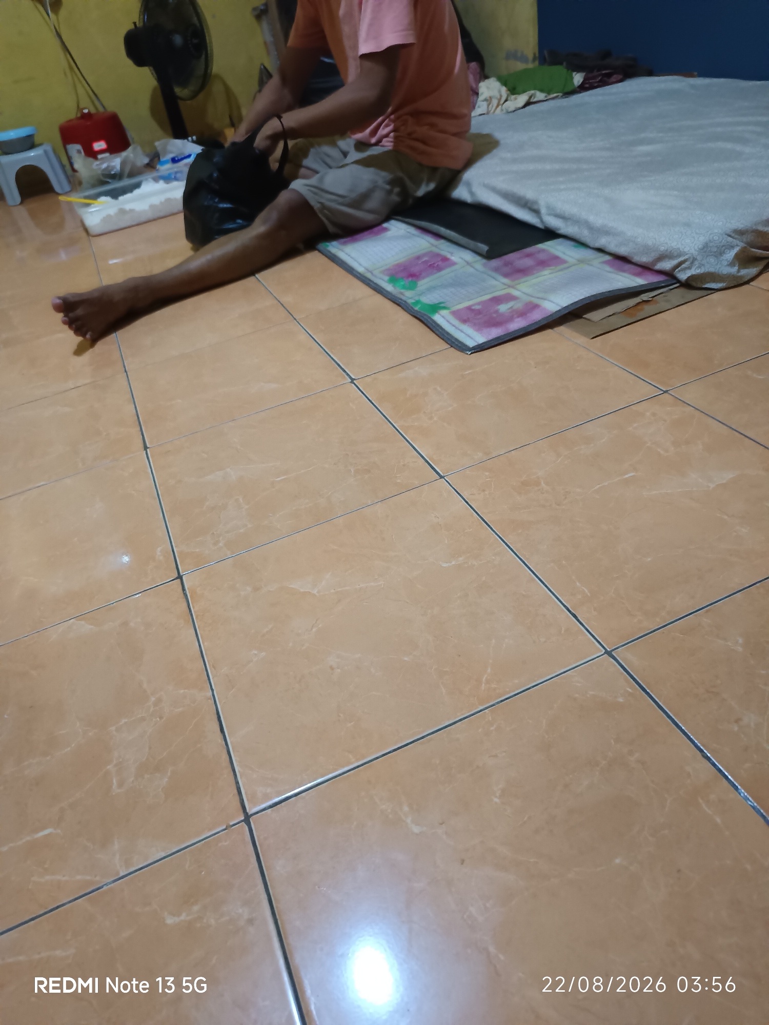 The image shows a person sitting on the floor in what appears to be a room. The person is wearing a pink t-shirt and grey shorts and is holding a black plastic bag. In front of the person, there is a mattress covered with a patterned sheet, and to the left, there are several objects including a fan, a red container, and a white bowl with white contents. The floor is tiled in a light brown color. A date and time stamp in the bottom right corner reads "22/08/2026 03:56", and in the bottom left corner, "REDMI Note 13 5G". The scene is likely a domestic environment. The specific location in Talaga Sari, Indonesia cannot be determined from the visual information provided.
