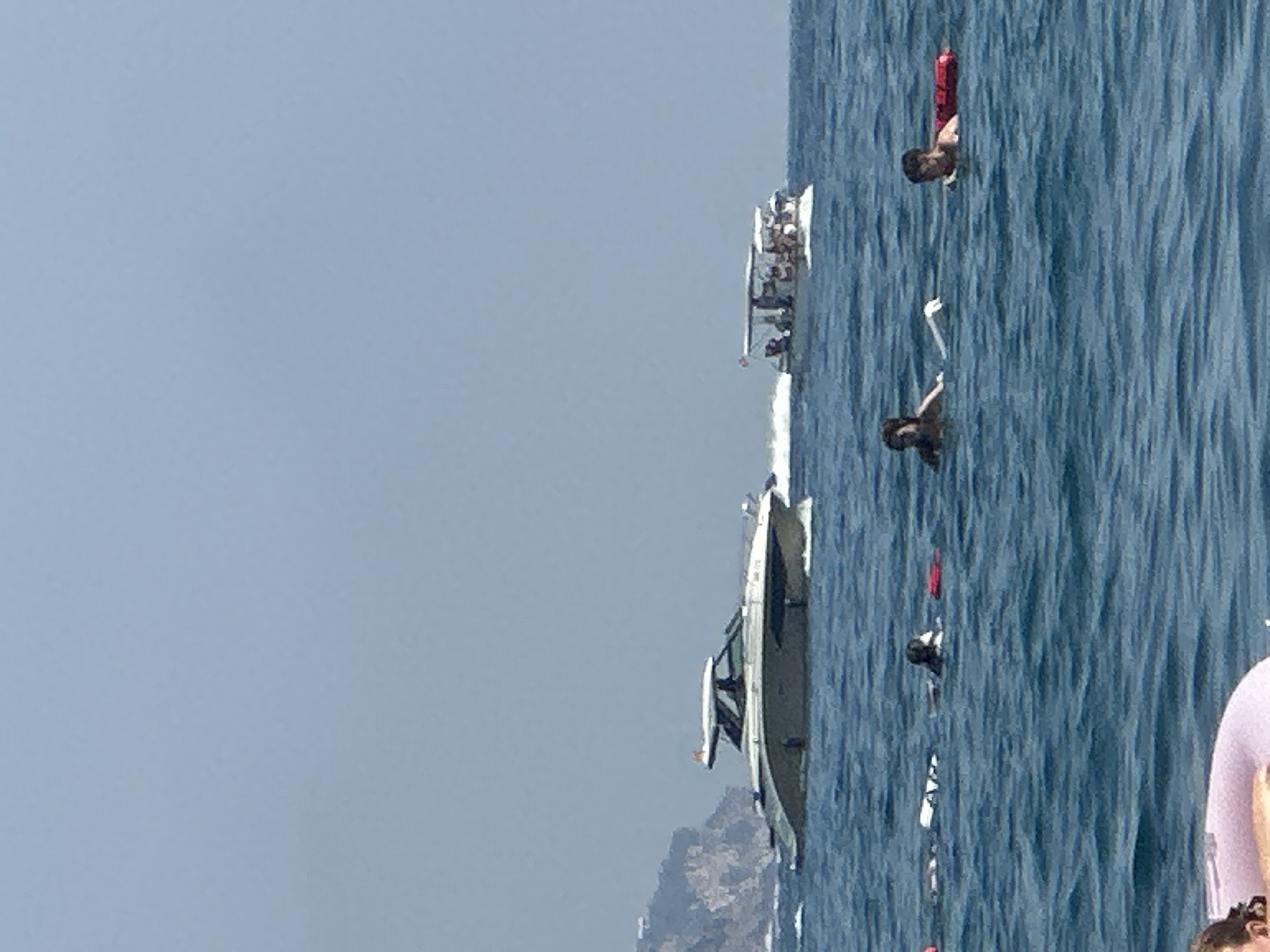 In the waters off the coast of Boreti, Montenegro, several individuals are enjoying recreational activities. A prominent modern yacht, light-colored with a dark hull accent and a white superstructure, is cruising in the mid-ground. Further in the distance, a smaller, open-top boat filled with passengers navigates the sea. In the foreground and mid-ground, several people are in the water. One person is on a pink inflatable ring, their upper body visible. Others are swimming or floating, with some wearing life vests. Red buoys are also visible in the water. The environment is a clear, blue sea under a pale sky, with a rocky, treed coastline in the far background.