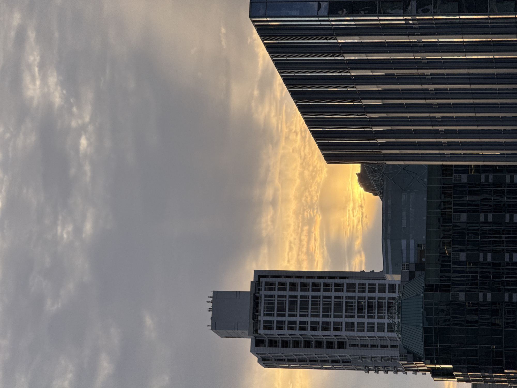 The image shows a cluster of modern buildings in Chicago, United States of America, set against a dramatic sky with the sun breaking through the clouds. In the foreground, a building with a glass facade and vertical silver fins stands prominently on the right. To its left, another building with a stepped, glass-covered roof is visible, and behind that, a tall, rectangular building with numerous windows dominates the left side of the frame. The sky above is filled with a mix of gray clouds and streaks of golden light emanating from the setting or rising sun, creating a striking contrast with the geometric lines of the architecture. There are no people or visible signs of activity. The overall impression is one of urban grandeur and the interplay of light and shadow on contemporary structures.