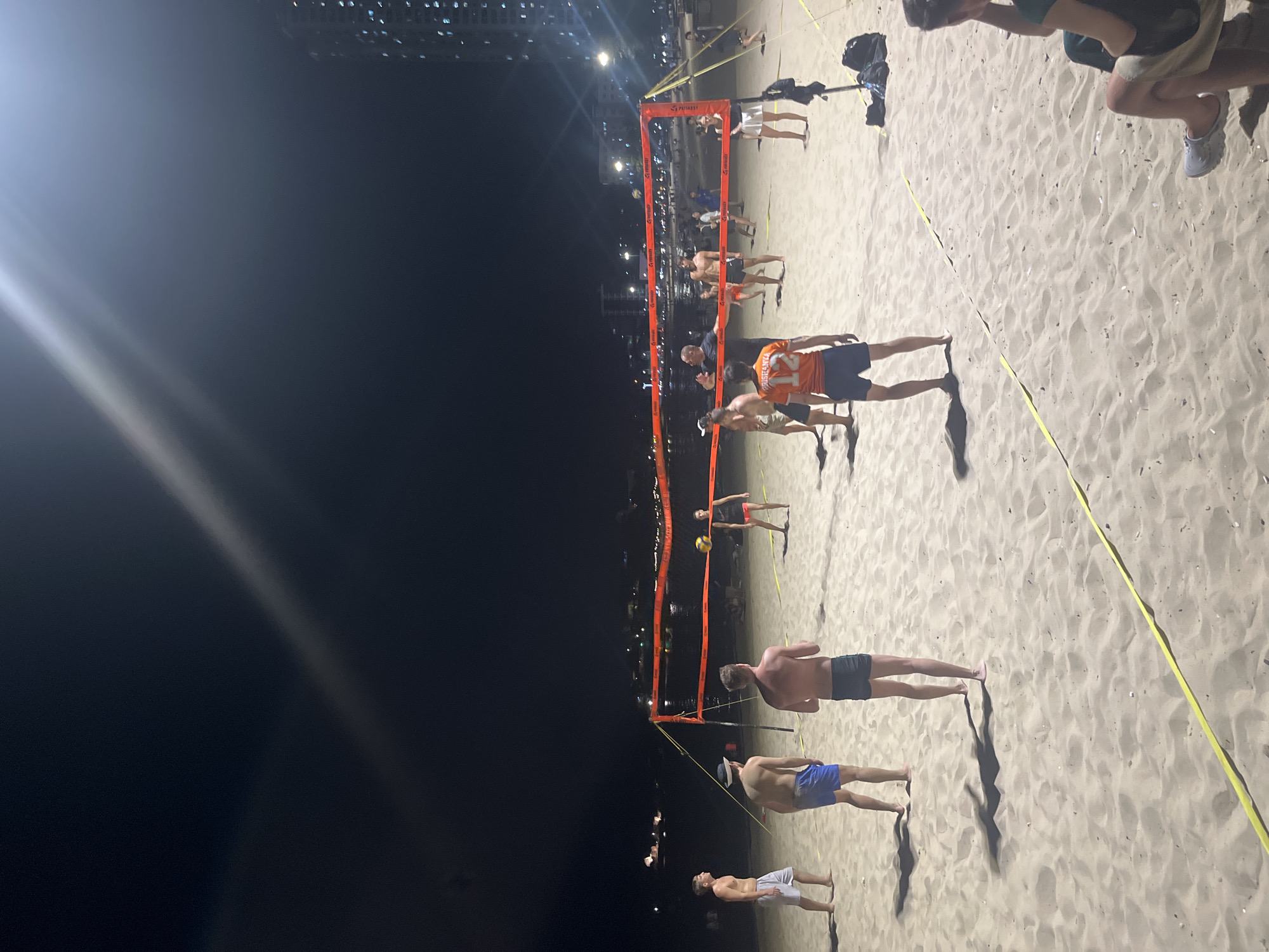 The image captures a nighttime beach volleyball game. Several shirtless men in swimwear are playing on a sand court defined by an orange net and yellow boundary lines. A volleyball is suspended in the air above the net. In the background, buildings with illuminated windows are visible against the dark sky. The scene is lit by artificial lights, creating long shadows on the sand. The city is Unknown, Unknown.