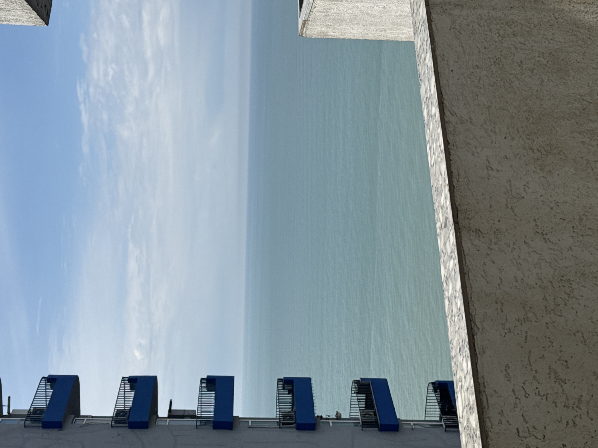 Captured from a high vantage point, the image presents an abstract composition where the division between sky, sea, and urban infrastructure blurs. A pale blue sky with scattered cloud formations occupies the upper left portion of the frame, contrasting with the expansive, opaque plane representing the sea. A section of a building with a textured facade dominates the right side, its neutral tone sharply juxtaposed against the blue elements. Below the horizon line, the roof of a building is visible, punctuated by a series of evenly spaced, blue-capped structures with metallic grating. These elements appear as functional architectural features, possibly related to ventilation or access. The location is likely Batumi, Georgia, suggested by the architectural style and coastal setting. The overall perspective creates a disorienting, almost architectural study.
