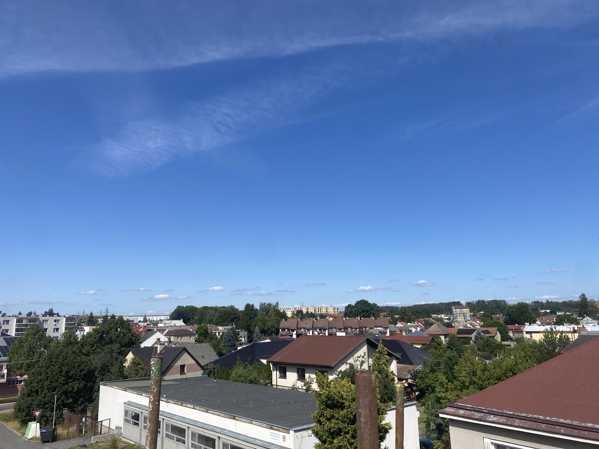 The image shows a panoramic view of a suburban area in Polička, Czechia, under a clear blue sky with wispy clouds. The foreground features the flat roof of a building with industrial pipes and some greenery. Beyond this, the scene unfolds into a dense collection of residential houses with diverse roofing materials, including dark grey, brown, and red tiles. Numerous trees are interspersed among the houses, creating a verdant landscape. In the background, taller apartment buildings and power lines are visible, indicating a mix of residential and utility infrastructure. The overall impression is of a peaceful, sun-drenched afternoon in a European town.