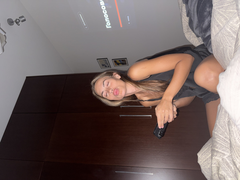 A woman in a dark tank top and shorts poses for the camera with pursed lips, holding a phone. She's positioned on a bed with a patterned duvet and a plush grey blanket. Behind her is a dark brown closet or wardrobe with silver handles. A television screen is visible on the ceiling, displaying text in a foreign language. The room is dimly lit, and two framed pictures adorn the wall above the closet.