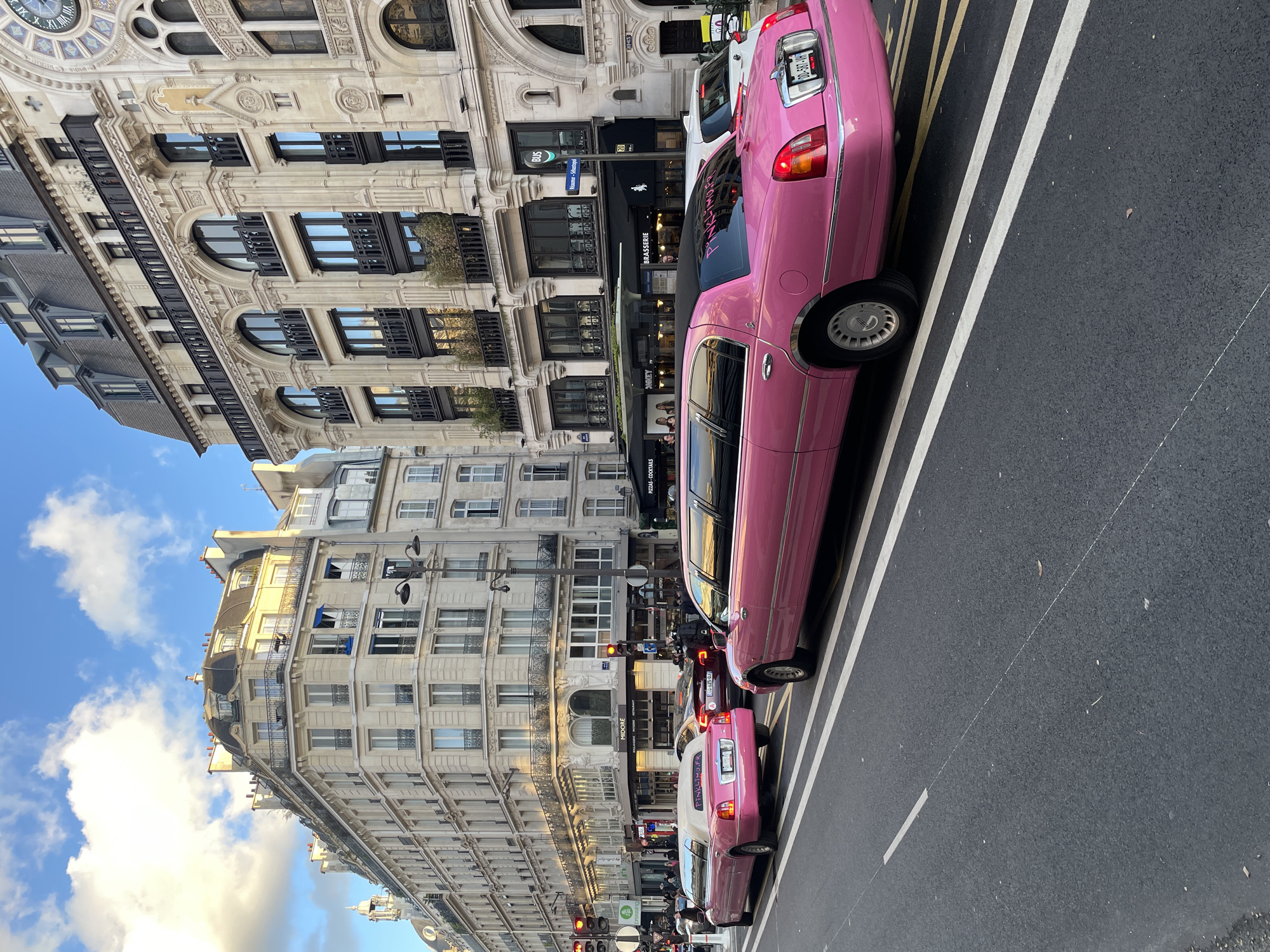 The image captures a street scene in Paris, France. Prominently featured are two pink limousines parked along the right side of the road, with white and yellow lane markings visible on the asphalt. The buildings lining the street showcase classic Parisian architecture, with multiple stories, ornate facades, and a mix of window styles. Street signs and traffic lights are present, indicating an active urban environment. A Starbucks can be seen in the background, and other cars and people are also visible in the distance. The sky is partially cloudy. The clock on the building in the background says that it is 11:00. A restaurant called "Brasserie" is also visible.