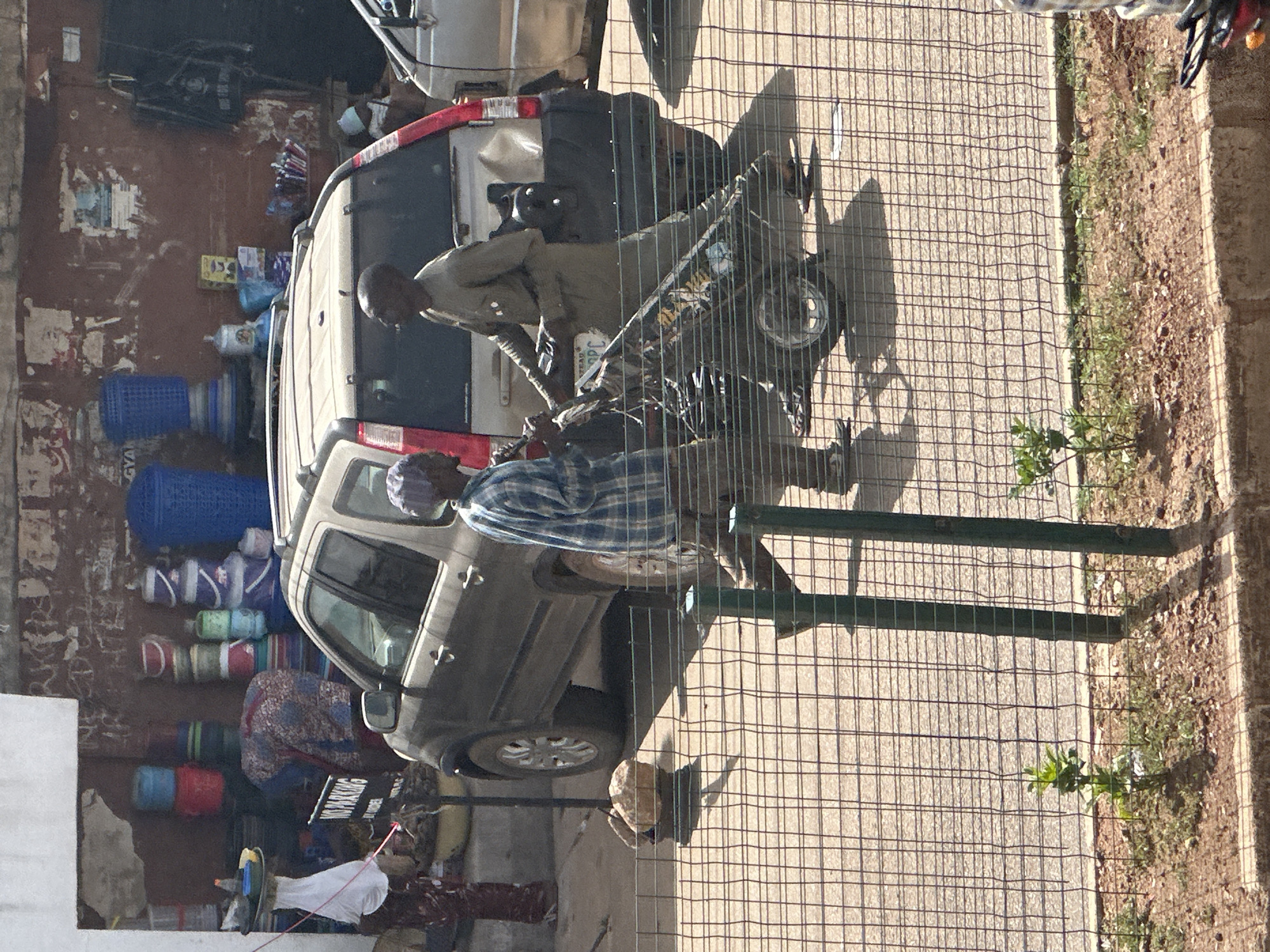The image shows an outdoor scene in what appears to be a busy area, possibly a market or street, in Bukuru, Nigeria. The perspective is from above, looking down at an angle.
The main subjects are two men and a vehicle. One man, wearing a khaki uniform, is crouched beside a light-colored SUV, his face partially obscured. Another man, wearing a blue and white checkered shirt, is also near the SUV. A motorcycle is parked or being handled next to the men and the SUV.
The scene is filled with various objects. Several large blue plastic baskets are stacked against a rough, reddish-brown wall on the left side of the image. Other colorful plastic containers and rolled-up items are also visible near the wall. The SUV appears to be parked, and there are some items on its roof and inside. A metal fence, painted green, runs across the foreground, partially obscuring the ground and the subjects. The ground is paved with concrete or asphalt and has some patches of dry grass and dirt along the edge where the fence meets the ground.
There is visible text in the image. A sign on the SUV reads "NO PARKING". Some faint writing or graffiti is present on the wall.
The time of day appears to be daytime, indicated by the shadows cast by the fence and the general lighting. The weather seems to be dry and sunny. The overall impression is one of daily activity and commerce, with people and goods in transit or for sale.