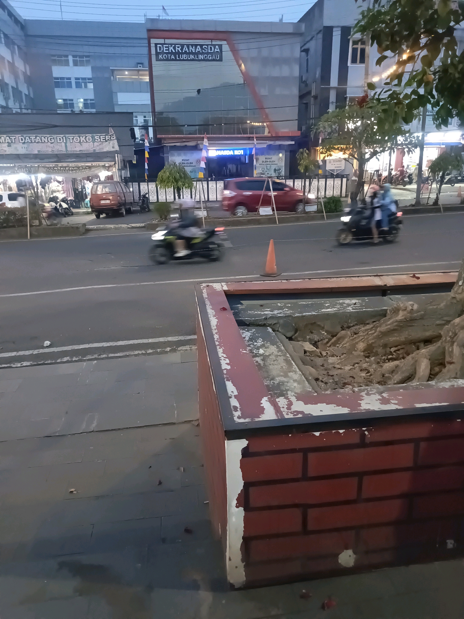 The image shows a street scene in Lubuklinggau, Indonesia, likely in the evening or early morning, given the low light conditions and artificial lighting from businesses. The main building in the background has signage that reads "DEKRANASDA KOTA LUBUKLINGGAU," indicating it is the Regional Crafts Council of Lubuklinggau City. Adjacent to this building is a store with an awning and merchandise visible, with text that includes "MAT DATANG DI TOKO SER" which translates to "Welcome to SER store."
In the foreground, there is a damaged brick planter box with exposed dirt and roots. The street is paved with asphalt and has a white lane marker. Several vehicles are present on the street, including a red car and two motorcycles with riders. One motorcycle is in motion, appearing blurred due to its speed, carrying two passengers. Another motorcycle is parked near a small red car. An orange traffic cone is placed on the side of the road. Trees are visible along the sidewalk, some with lights strung on them. The overall environment suggests a commercial area of the city.