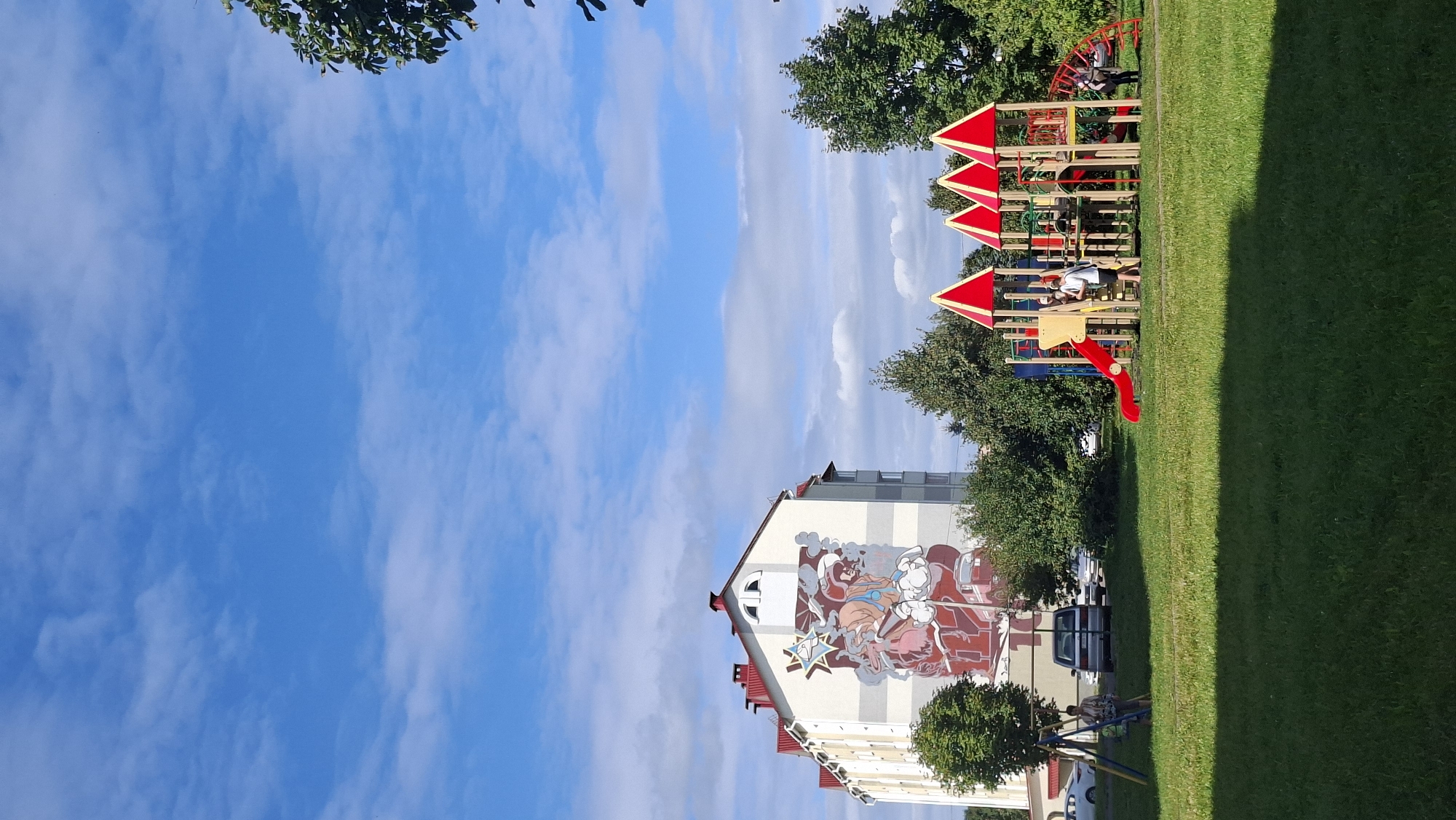 In Myadzyel, Belarus, a multi-story residential building with a large mural on its side stands next to a playground. The mural depicts a stylized image of a fire truck and figures in red and white. The playground features a red slide, climbing structures, and red triangular roof elements. Several people are visible on the playground, including a woman near a swing set and children on the play structures. Cars are parked in the background near the building. The scene is set against a bright blue sky with scattered white clouds, and lush green grass covers the ground.