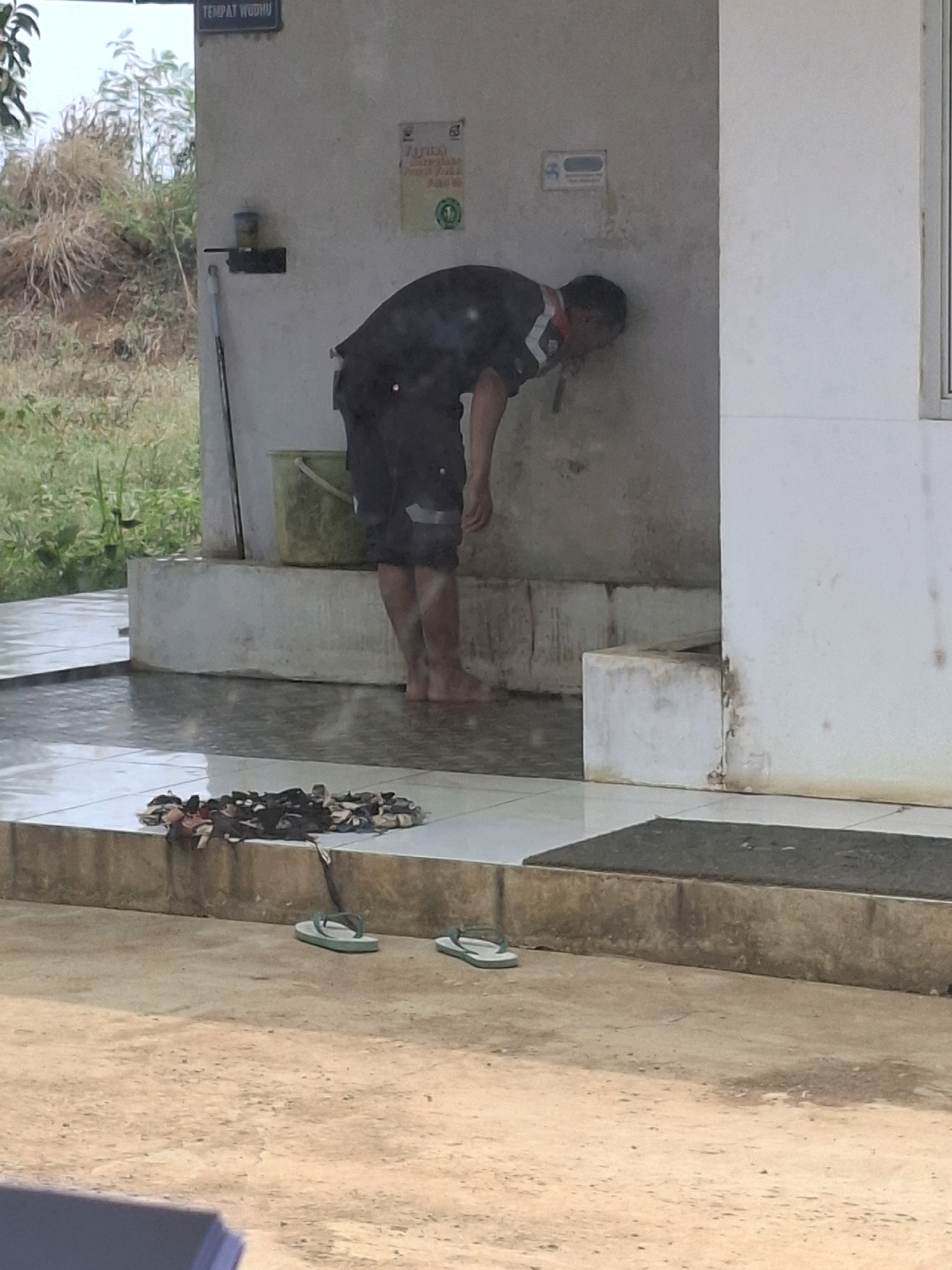 In Ciheulang Tonggoh, Indonesia, a man in a dark uniform with reflective stripes on the sleeves and shorts is performing ablutions. He is bending over, with his face close to a rough, gray wall, and appears to be washing his face or mouth. Water is visible on the tiled floor in front of him, suggesting a washing area. To his left, a light green bucket sits on a raised platform. Behind him, an area with dry vegetation and trees can be seen. In the foreground, several pairs of flip-flops are scattered on the ground, and a pile of clothing lies on a ledge. A dark mat is placed on a concrete step. A sign on the wall above the man's head reads "TEMPAT WUDHU," which translates to "ablution place" in Indonesian.