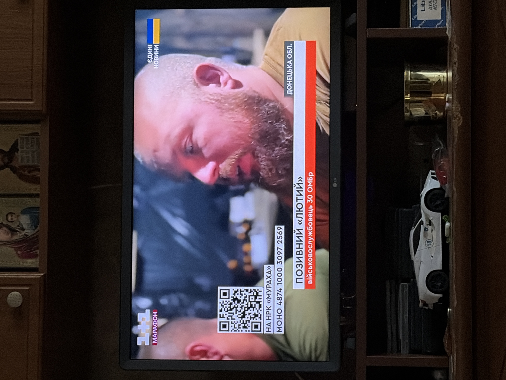 The image shows a television screen displaying a news broadcast. On the left side of the screen, the "1+1" logo is visible, followed by the word "МАРАФОН" in red. Below this, a QR code is displayed, with text underneath reading "НА НРК «МУРАХА»" and a phone number "МОНО 4874 1000 3097 2569". The center of the screen features a man with a beard and light-colored hair. A banner at the bottom of the screen shows text in Ukrainian: "ПОЗИВНИЙ «ЛЮТИЙ»" and "ВІЙСЬКОВОСЛУЖБОВЕЦЬ 30 ОМБР". To the right of this, it reads "ДОНЕЦЬКА ОБЛ.". In the top right corner of the screen, the Ukrainian flag colors and the words "ЄДИHI НОВИНИ" are visible. The television itself is black and has an LG logo at the bottom center. The television is placed on a dark brown shelf. Below the shelf, a white toy sports car with a circular emblem featuring the number 18 is visible. To the right of the car is a cylindrical brass object. Further to the right, a box with blue and white markings, partially obscured, is visible. Above the television, there are two religious icons displayed on a wooden shelf.