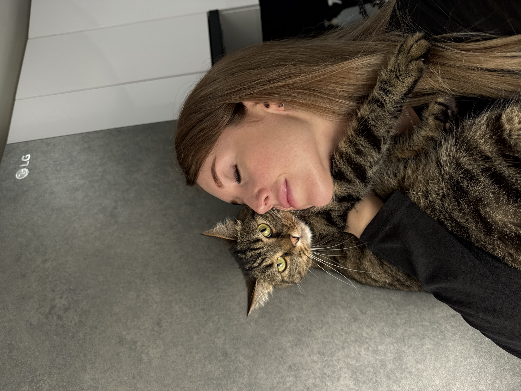 In a well-lit, indoor setting, a young woman with long, blonde hair and closed eyes is embracing a tabby cat. The woman is wearing a black long-sleeved shirt. Her head is tilted, resting against the cat's head. The cat, with striking green eyes, appears alert and is looking directly at the camera. The background consists of a textured grey wall to the left and a white, paneled surface to the right, where an "LG" logo is visible. This scene is not identifiable as being in the city of Chaikovsky, Russia.