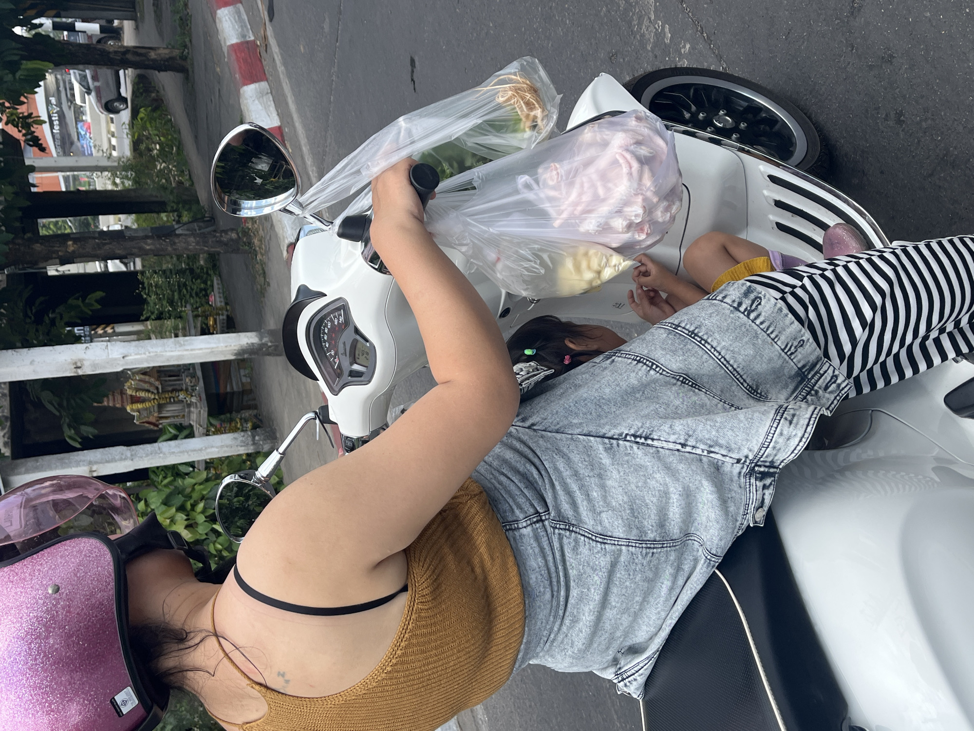 Captured in Chiang Mai City Municipality, Thailand, the image shows a woman on a white scooter. She is wearing a pink helmet, a mustard-colored top, and a faded denim jacket. A child wearing black and white striped pants is seated on the scooter with her. Plastic bags filled with food items hang from the scooter's handlebars. The background includes a street, trees, and a building in the distance.