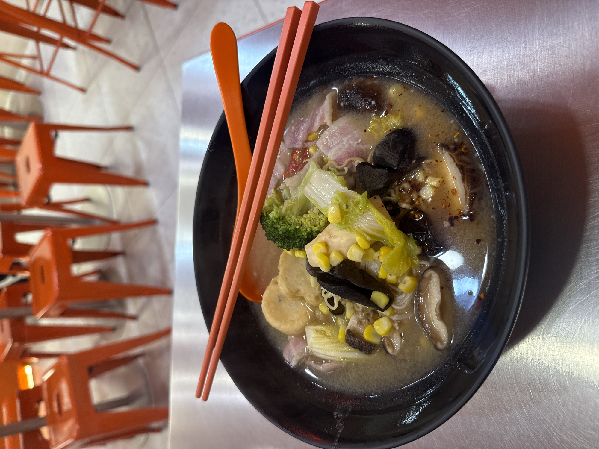 The image shows a bowl of ramen with broccoli, cabbage, corn, mushrooms, and sliced pork belly. The bowl is black and there is an orange spoon and chopsticks resting on the rim. The bowl is placed on a silver metal table with several orange metal chairs stacked to the left. The image was taken in Bangkok, Thailand.