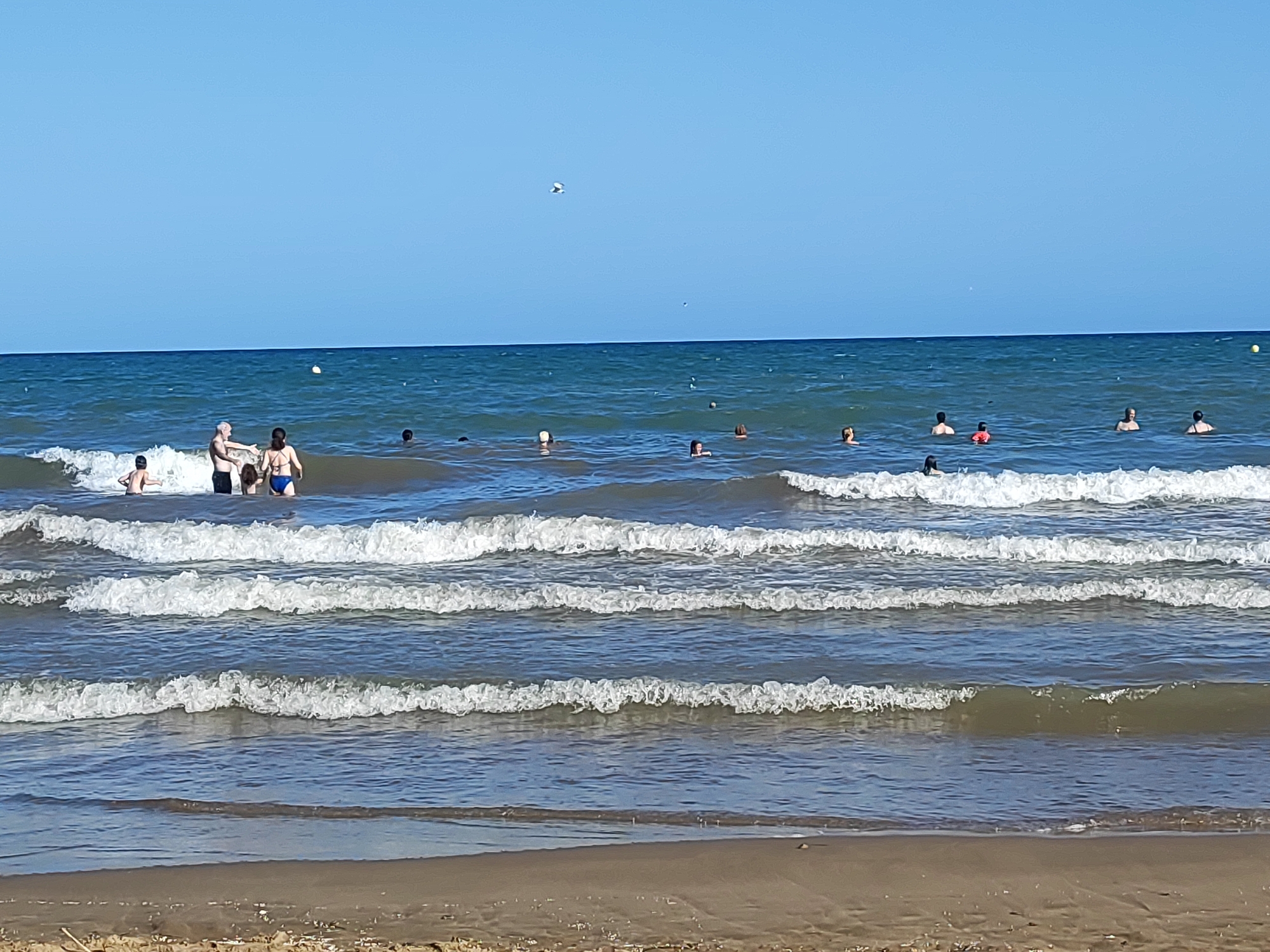 In Playa, Spain, a group of people enjoy the ocean. The scene is dominated by the blue expanse of the sky and the turquoise water, with gentle waves breaking on the sandy shore. Several individuals are visible in the water, some swimming, others standing. A man in black shorts and a woman in a blue bikini are near the shore, engaged in conversation. Further out, a boy in a red vest is visible in the water. The sunlight creates a bright, clear atmosphere, ideal for a day at the beach.