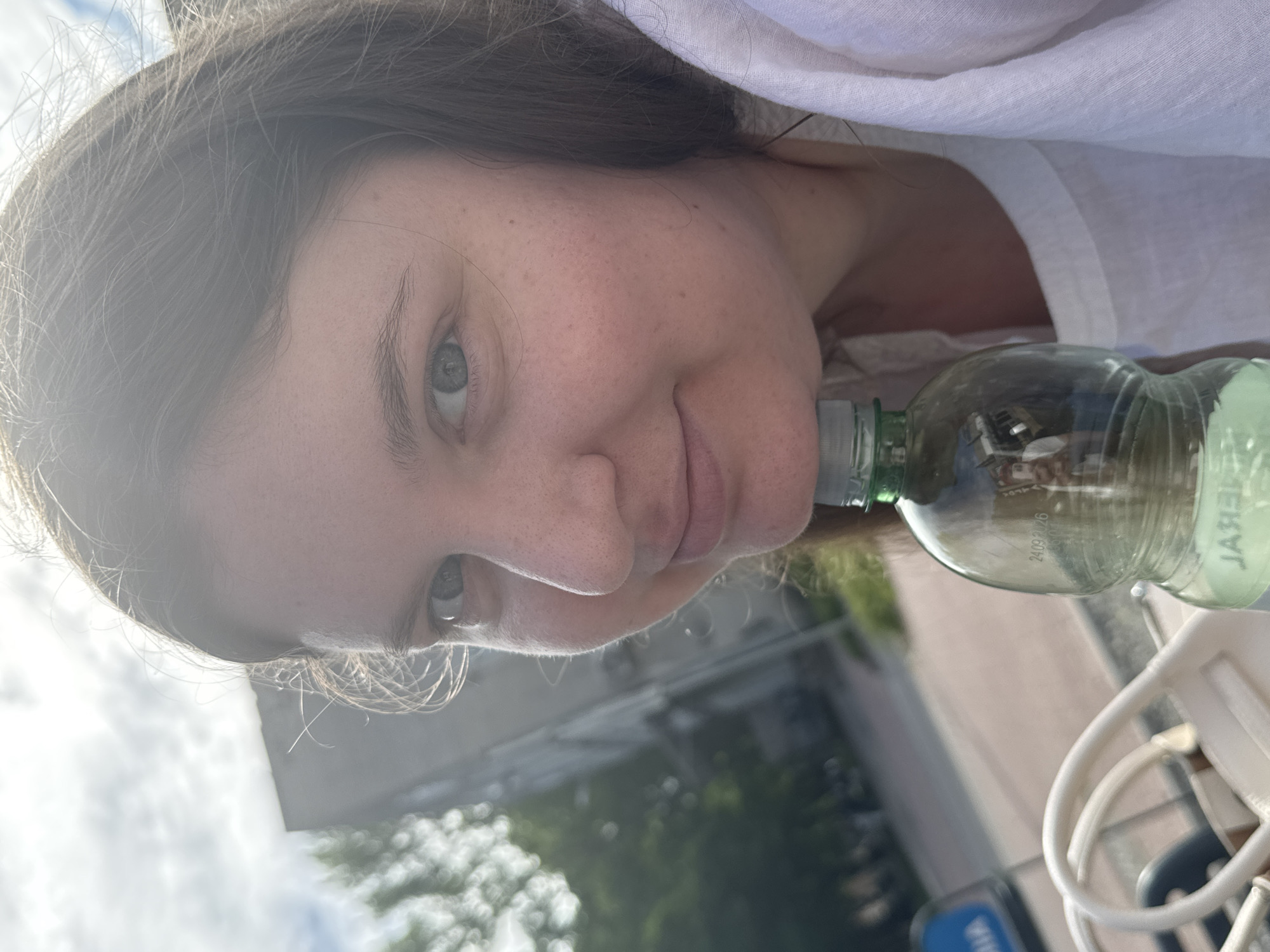 A close-up, selfie-style photograph shows a young woman with shoulder-length brown hair and fair skin, looking towards the camera. Her left eye is clearly visible, along with her nose and mouth. She is resting her chin on the neck of a clear plastic water bottle, which is positioned vertically in front of her. The bottle has a green cap and appears to contain a light green liquid. The woman is wearing a white garment, possibly a shirt or blouse, with a textured fabric visible on the right side of the image.
The background is blurred, suggesting an outdoor setting. To the left and slightly behind the woman, a building with a grey facade and several windows can be seen. Green foliage, likely trees, is also visible in the background, some of which is out of focus and creates a bokeh effect. The sky is visible at the top of the image, with some clouds and sunlight filtering through, creating a lens flare effect in the upper right quadrant. A portion of what appears to be a white bag with curved handles is visible in the lower left corner of the image. The ground or pavement in the middle ground appears to be a light-colored surface with some texture. A blue sign with white lettering is partially visible in the far left background. The lighting suggests it is daytime, likely late afternoon or early morning due to the angle of the sun.
The scene appears to be set in an urban environment, possibly a park or a street with pedestrian areas, as indicated by the paved ground and the presence of a building. The overall impression is a casual, spontaneous moment captured outdoors. The timestamp on the bottle reads "24.09.2026", suggesting it is a product with an expiration date.
While the exact location within Tallinn, Estonia, cannot be definitively determined from the visual information alone, the presence of a building with the architectural style visible and the general urban outdoor setting are consistent with locations found within the city.