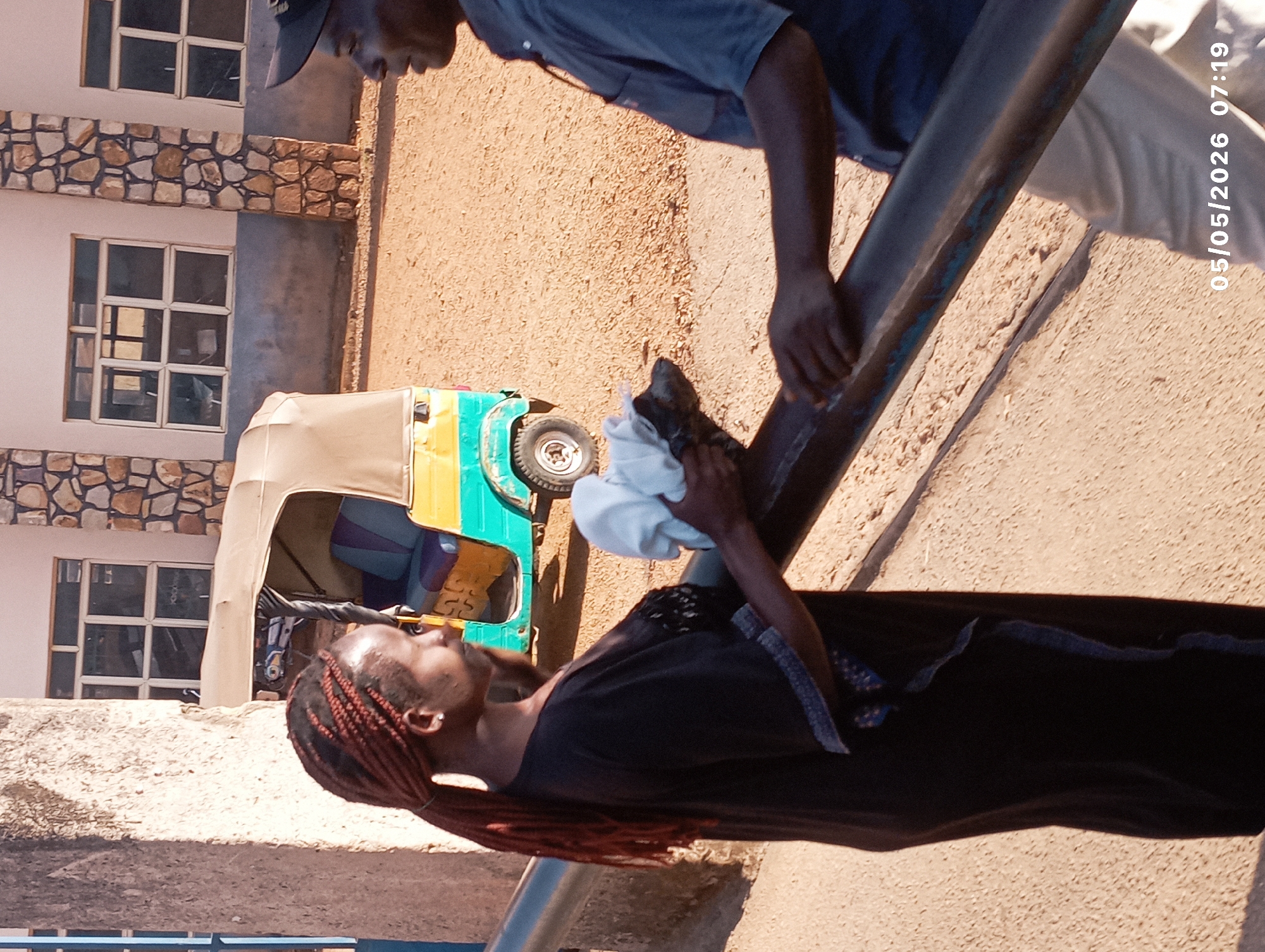 Two people stand near a blue railing; one person wearing a black shirt and brown braids holds a white cloth and looks upward, while the other, wearing a blue shirt and baseball cap, places his hand on the railing. A yellow and blue auto-rickshaw is parked nearby. The background includes a building with windows and stone details, a dirt area, and the date "05/05/2026 07:19." The city is assumed to be Jos, Nigeria.
