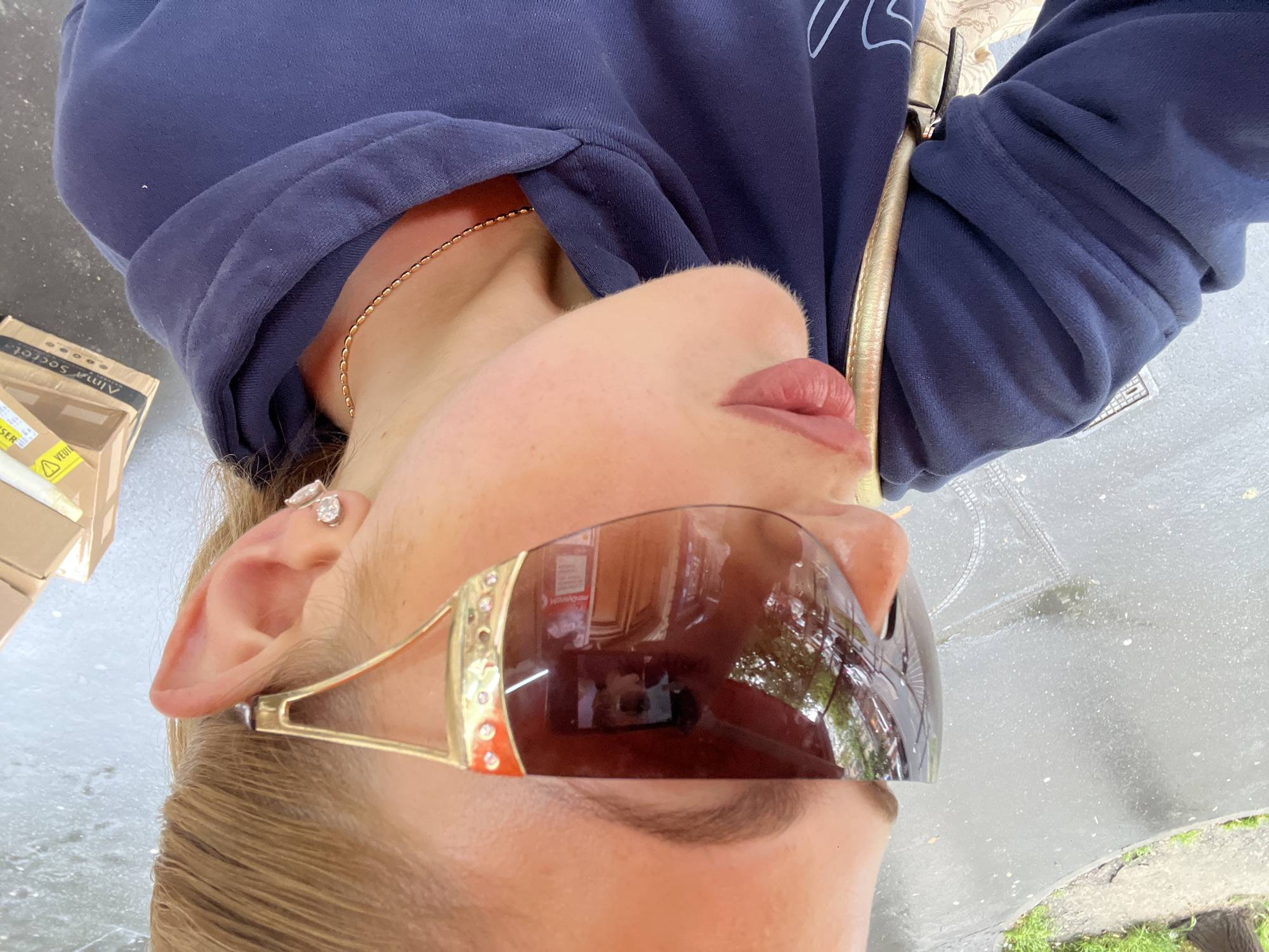 A woman wears sunglasses with a gold frame and red accents, her blond hair pulled back. She has a gold chain necklace and earrings. She is wearing a navy blue hoodie. The reflection in her sunglasses shows an interior with a white-painted ceiling, a red sign, and possibly people. A box with "Alma Secret" is visible to the left. The image seems to be taken on a wet sidewalk in Paris, France, as hinted by the reflection in the glasses.