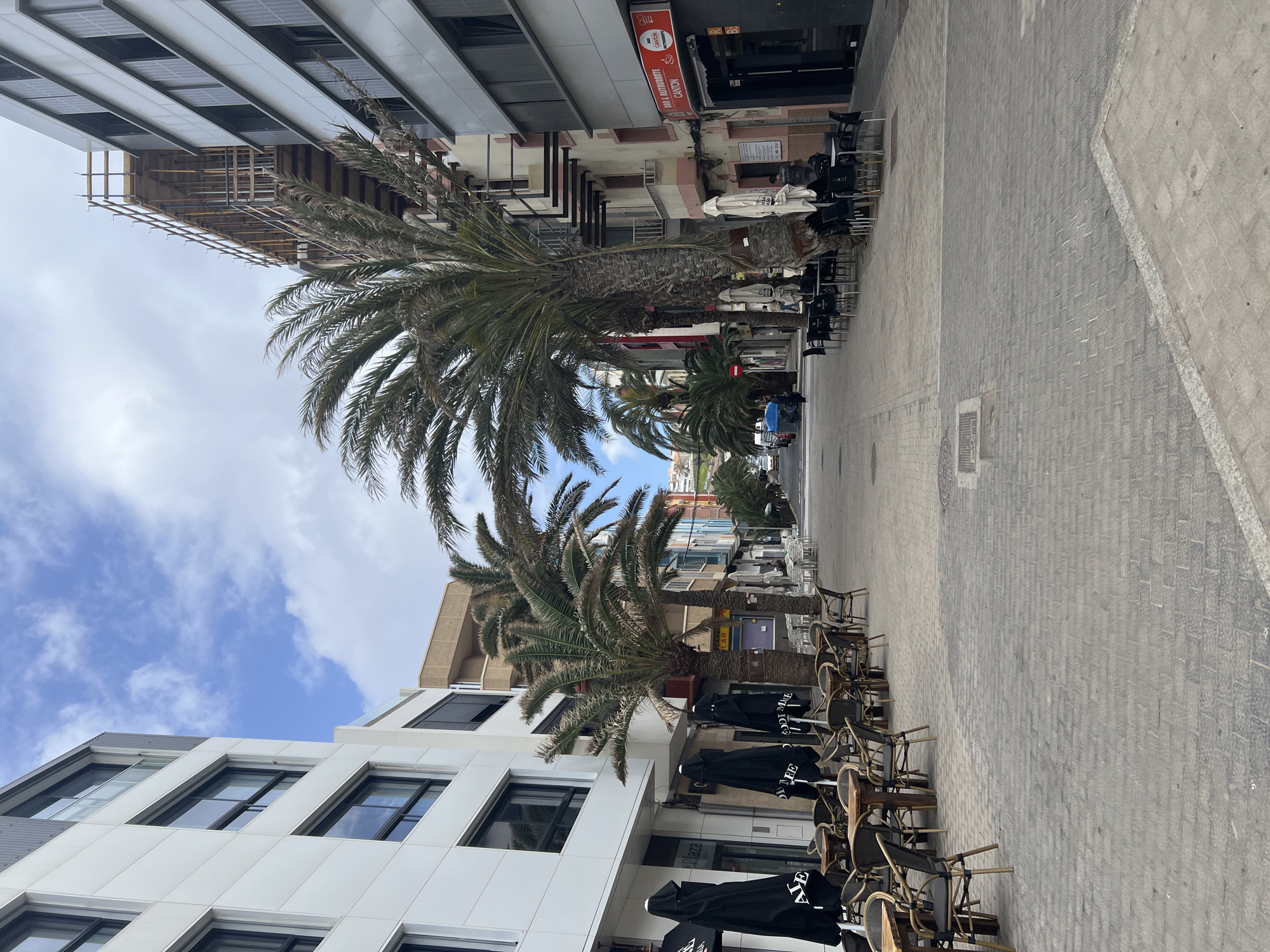 The image captures a street scene in Las Palmas De Gran Canaria, Spain. The street, paved with gray bricks, is lined with buildings of various heights and architectural styles. Palm trees are planted along the street, their fronds reaching towards the sky. Outdoor seating areas are set up in front of the buildings, with tables and chairs arranged under black umbrellas. The sky is blue with scattered white clouds. There are no people visible in the image.