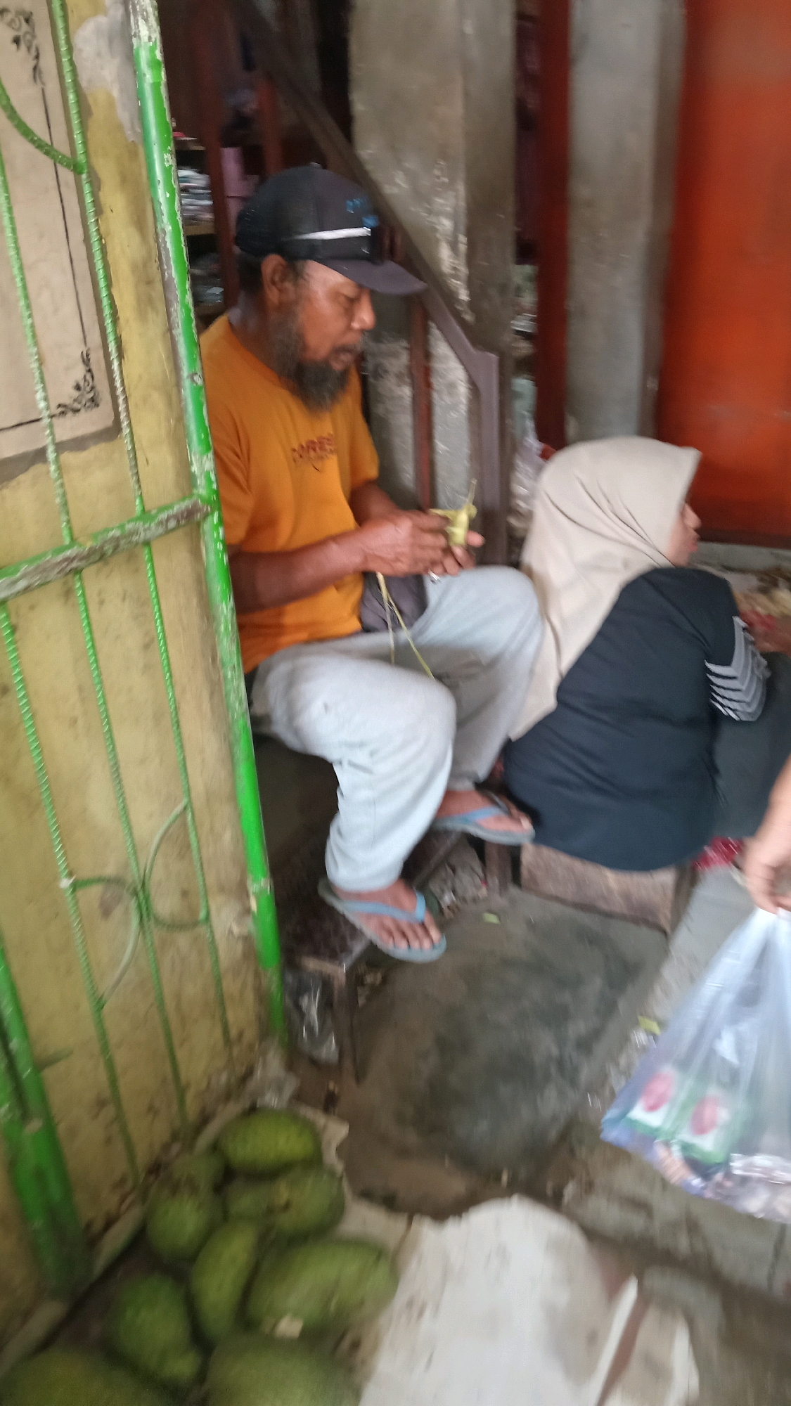In Kunduran, Indonesia, a man wearing a black cap and an orange t-shirt sits on a wooden stool, crafting with his hands. He has a beard and appears to be weaving or braiding something with palm leaves. Beside him, a person wearing a light brown hijab and a black outfit sits on another stool. In the foreground, a pile of green fruits, possibly mangoes or avocados, rests on the ground. To the right, a transparent plastic bag contains two small cartons with red and white labels, possibly juice boxes or beverages. The scene appears to be in an outdoor or semi-outdoor setting, possibly a market stall or roadside vendor, with a green-painted metal gate visible on the left and rough concrete or plastered walls in the background.