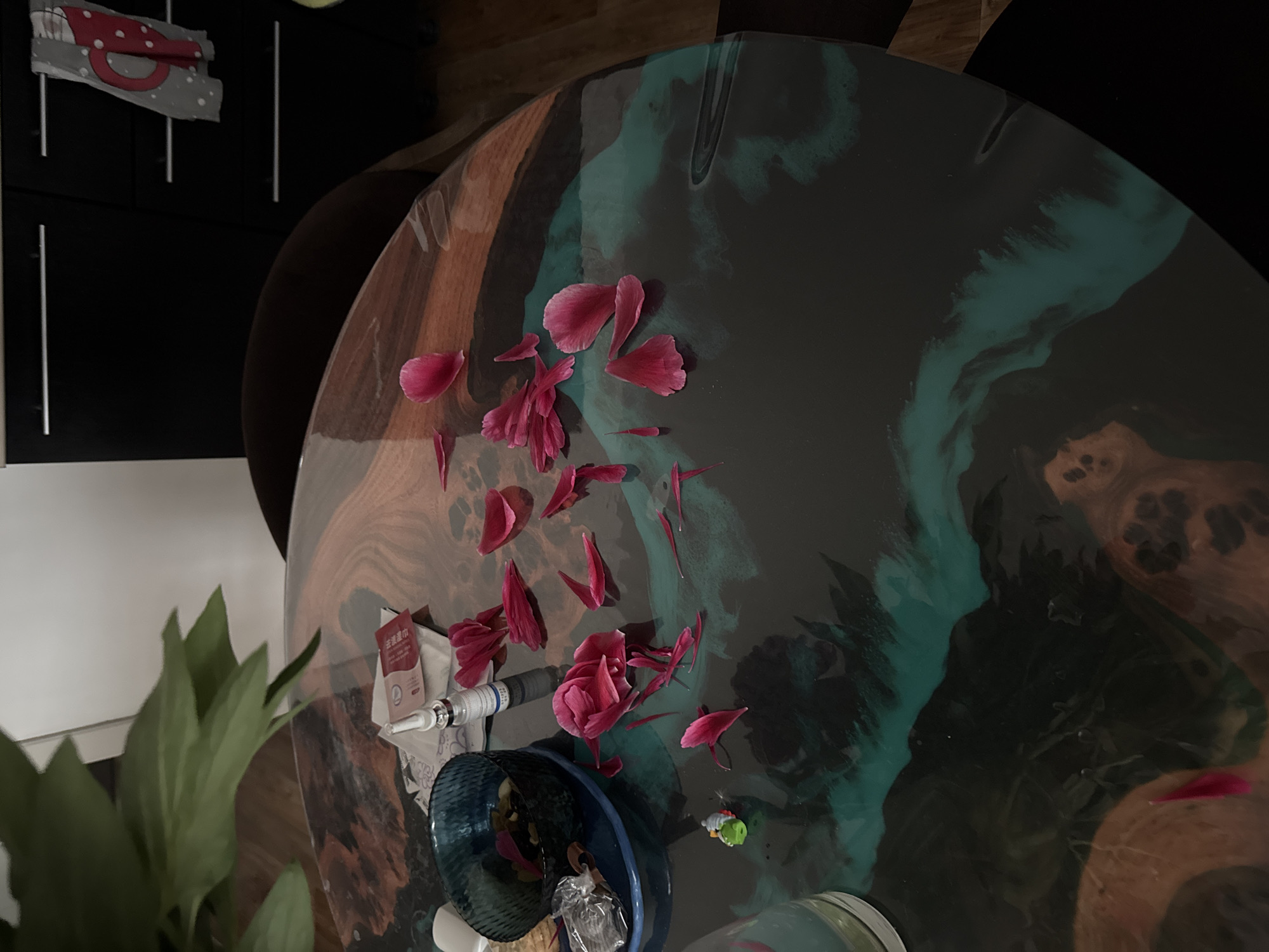 A high-angle shot depicts an abstractly painted round table featuring brown, black, and teal tones. Scattered atop the table are loose, pink flower petals. Positioned towards the lower left corner of the table are a blue glass bowl containing unidentifiable objects, a medical syringe, and a white box with red Chinese lettering. In the background, one can see dark furniture and a green leafy plant. Based on visual cues, it is not possible to confirm if the image was taken in Perm, Russia.