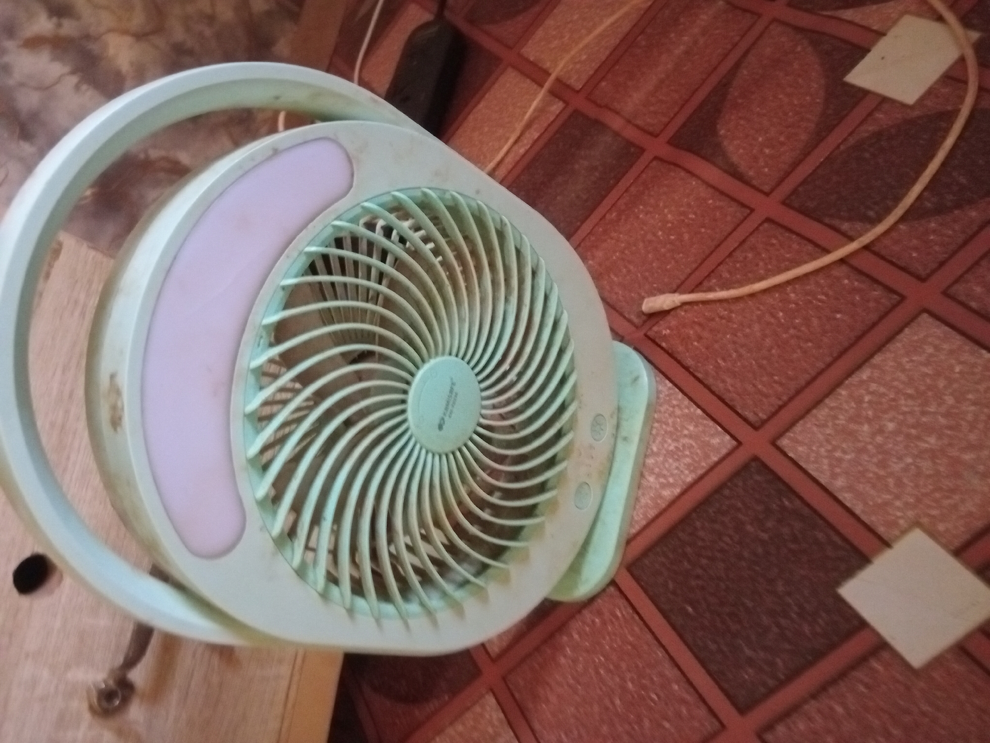 A tabletop fan with a light green frame and a pink accent is positioned in front of a background of red and brown tiled flooring. The fan has a circular grill and a handle, suggesting its portability. A power cord extends from the fan towards the upper right. The scene appears to be indoors, with a possible section of furniture visible to the left. There are no people visible. The location of Shagamu, Nigeria is not verifiable in this image.