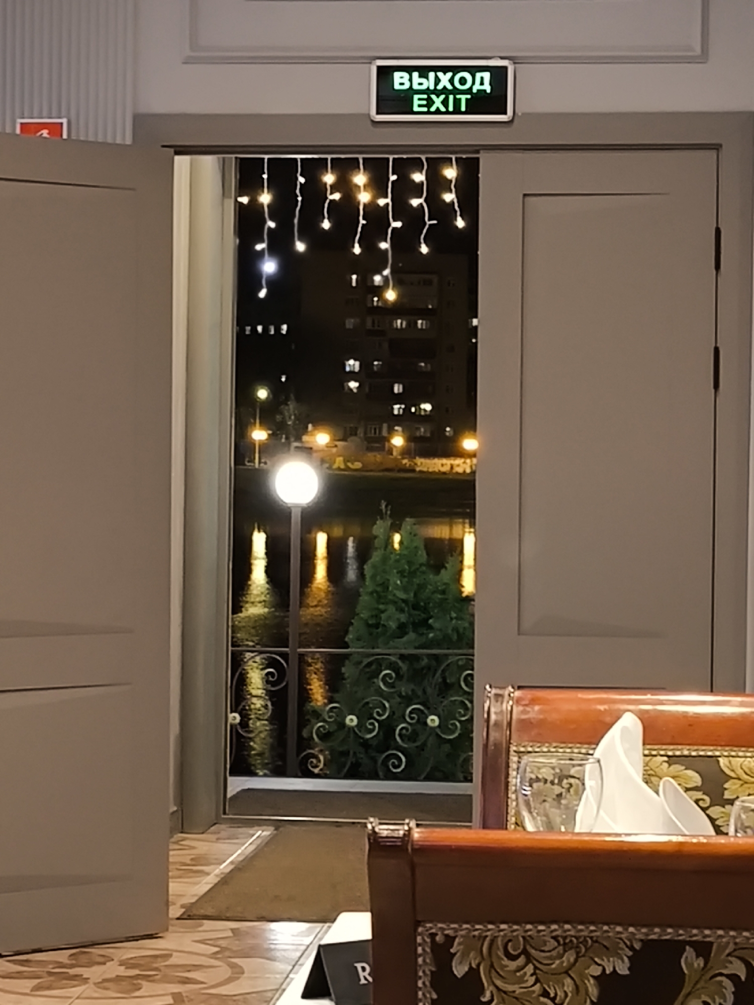 The image captures an indoor view, likely from a restaurant or cafe, looking out through an open doorway at night. In the immediate foreground, part of an ornate wooden chair with dark and gold patterned upholstery is visible, along with a white folded napkin and a glass on a table, suggesting a dining setting. The floor inside is tiled with a light brown and off-white pattern.
Through the doorway, a nighttime exterior scene unfolds. A calm body of water, possibly a river or lake, reflects the glowing artificial lights. A tall streetlight with a round white lamp stands prominently near the water's edge, its light reflecting brightly. In the distance, across the water, apartment buildings with illuminated windows are visible against the dark sky, indicating an urban or suburban environment consistent with Khimki, Russia. Decorative string lights with small, warm bulbs are strung across the top of the doorway, casting a gentle glow. A dark, ornate metal railing runs along the outdoor perimeter, and some green foliage, possibly a shrub or small tree, is seen behind it. A brown doormat is placed just outside the doorway.
Mounted above the doorway is a bright green exit sign with the words "ВЫХОД" (Russian for Exit) and "EXIT" displayed beneath it. To the far left, a small, blurry red sign is partially visible on the wall. No people are present in the image, lending a peaceful and serene atmosphere to the scene. A partial letter "R" can be seen on a black stand on the table in the bottom right corner, possibly a menu holder or reservation marker. The overall impression is one of a quiet evening view from a comfortable establishment.