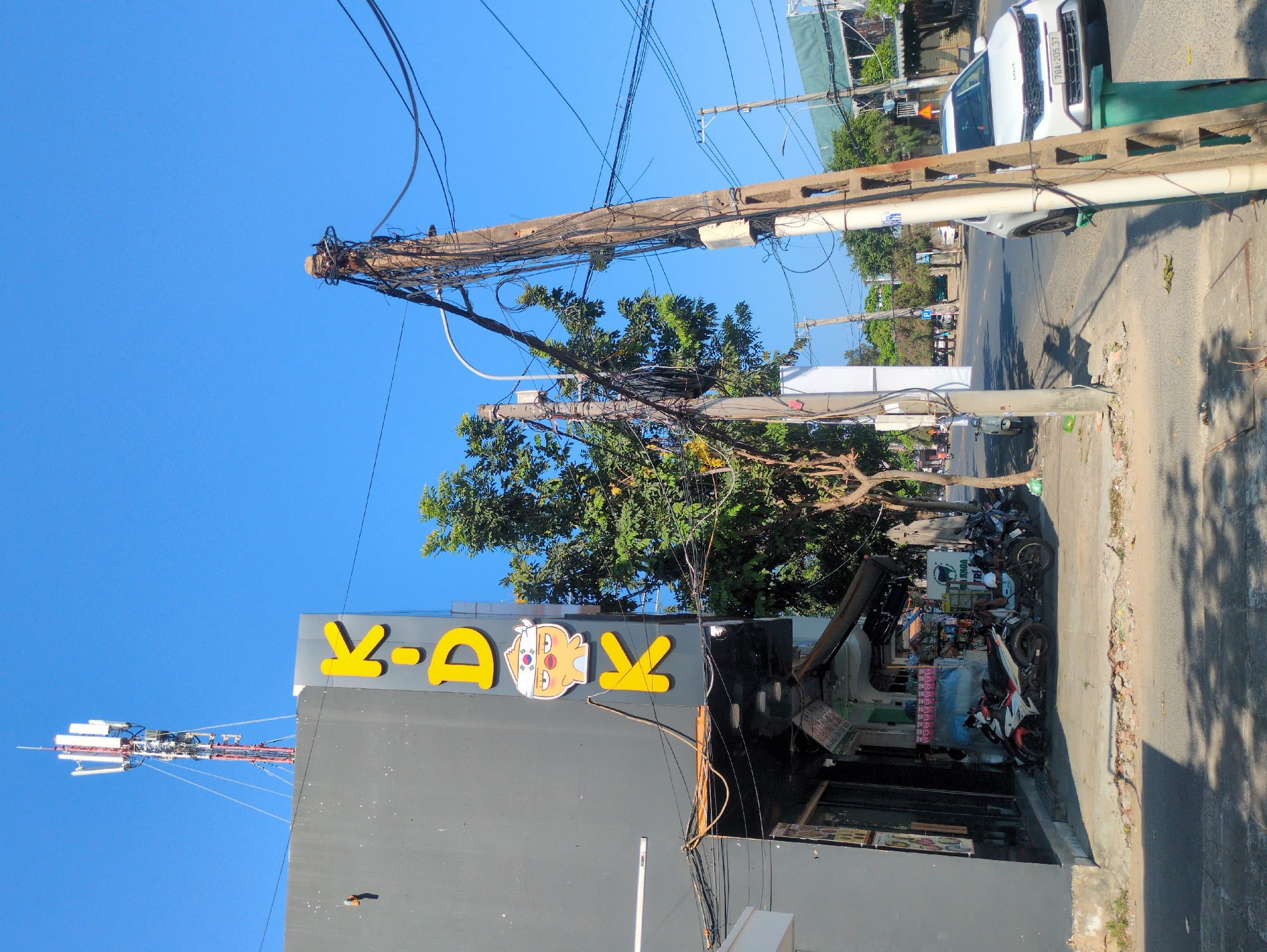 A street scene in Tuy Hòa, Vietnam features a gray building with a yellow "K-A-OK" sign and a character on its roof. A cell tower extends above the building. Power lines and utility poles are heavily bundled with wires. Several parked motorbikes are on the sidewalk in front of a commercial establishment. A white KIA car is parked on the street further down the road. The sky is clear and blue.