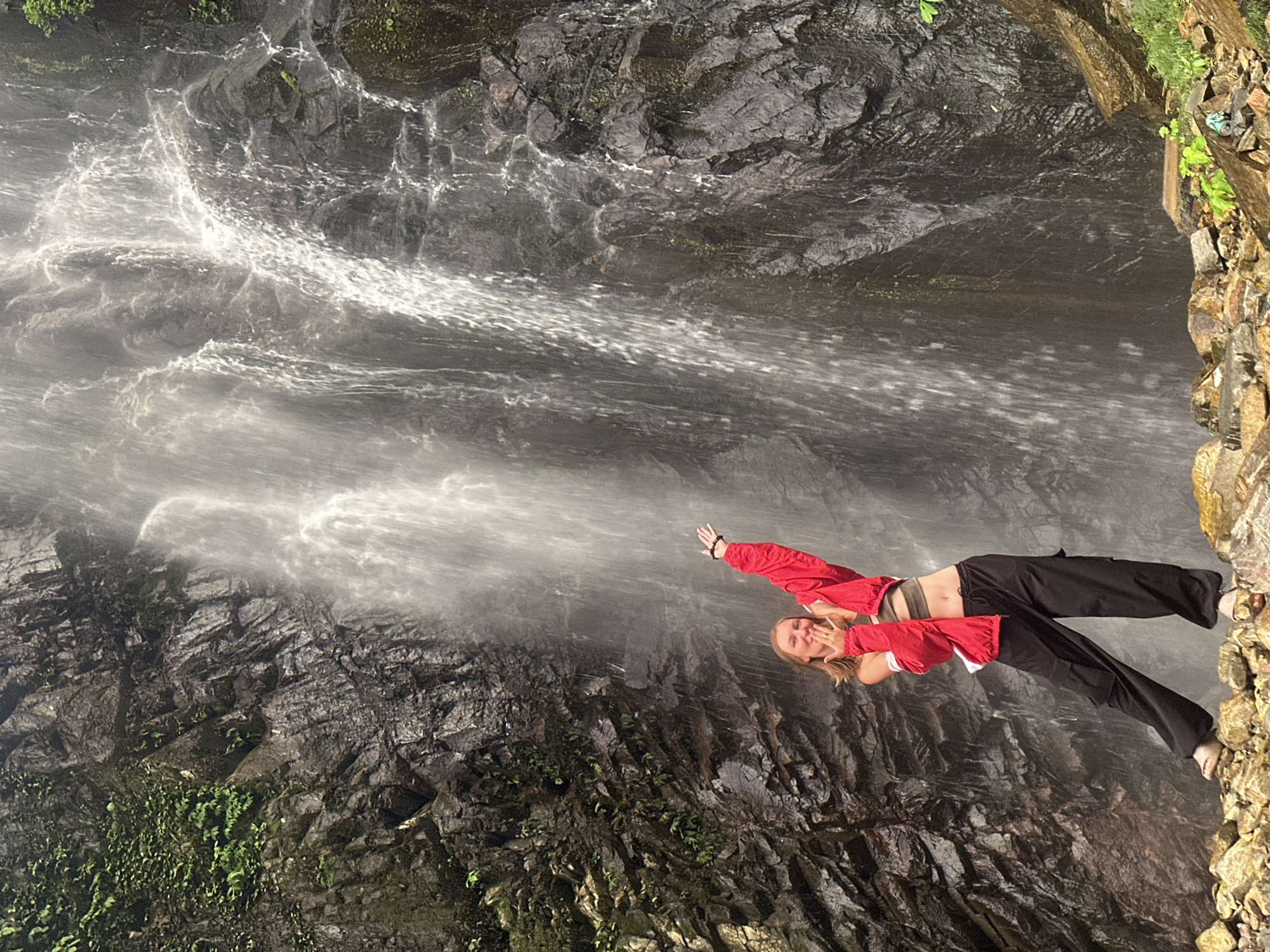 A full-body outdoor shot captures a young Caucasian woman standing on rocks at the base of a tall waterfall in Tu Le, Vietnam. The woman, with long blonde hair, is positioned slightly to the left of the frame's center, facing the camera with a smiling expression and her mouth partially open, with her left hand brought towards her mouth in a gesture, and her right arm raised with an open hand. She wears a dark olive-green crop top, a bright red long-sleeved jacket worn open and loosely over her shoulders, and baggy black cargo pants. On her feet are light-colored open-toed sandals. Her posture is dynamic, with her left leg extended forward and her right leg bent at the knee, giving the impression of movement or an active pose.
Behind her, the impressive waterfall dominates the scene, cascading down a rugged, dark grey rock face that is partially covered in lush green moss and small plants, particularly on the left side of the frame. The water flow appears strong and wide, creating a misty atmosphere. The rocks beneath the woman are irregular, angular, and vary in size and color, ranging from dark greys to lighter browns and oranges, suggesting a natural, untamed environment. Scattered green foliage is visible among the rocks in the lower right foreground. The lighting suggests an overcast or shaded condition, highlighting the water's movement and the textural details of the rocks.