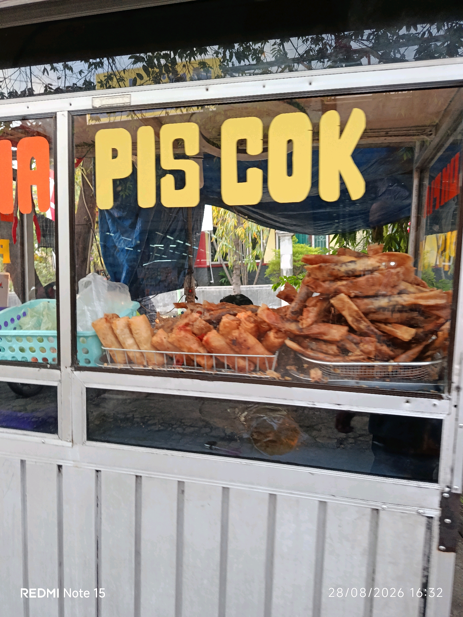 The image shows a food cart with a glass display case. Yellow lettering on the glass reads "PIS COK". Inside the display case, there are two metal baskets filled with fried snacks. One basket contains what appear to be spring rolls, while the other is filled with a pile of long, thin fried items, possibly fried fish or squid. To the left of the display, a light blue plastic basket holds some white plastic bags. The cart itself is white metal with vertical slats. The background shows some greenery and buildings, suggesting an outdoor setting in a neighborhood. The bottom of the image has a timestamp overlay that reads "28/08/2026 16:32".