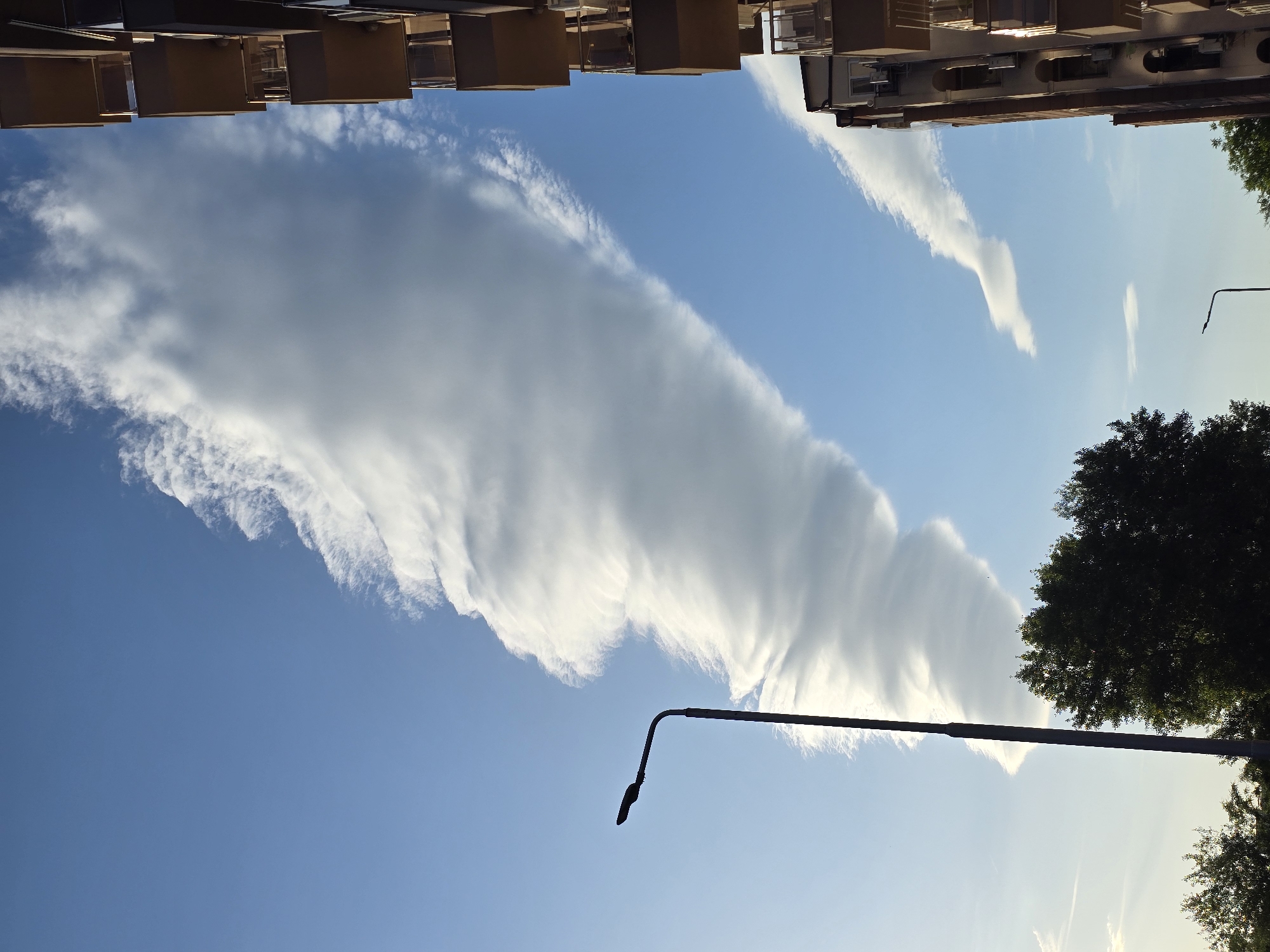The image depicts a low-angle view towards the sky, framed by urban elements. At the top, a section of a building with overhanging balconies is visible. Dominating the sky is a large, linear cloud formation with a pronounced wave-like structure, appearing white against a blue sky. A street lamp post extends across the lower part of the frame, partially obscuring the clouds. To the right, trees are visible, suggesting a park or green area nearby. The image is taken in the city of Unknown, Unknown.