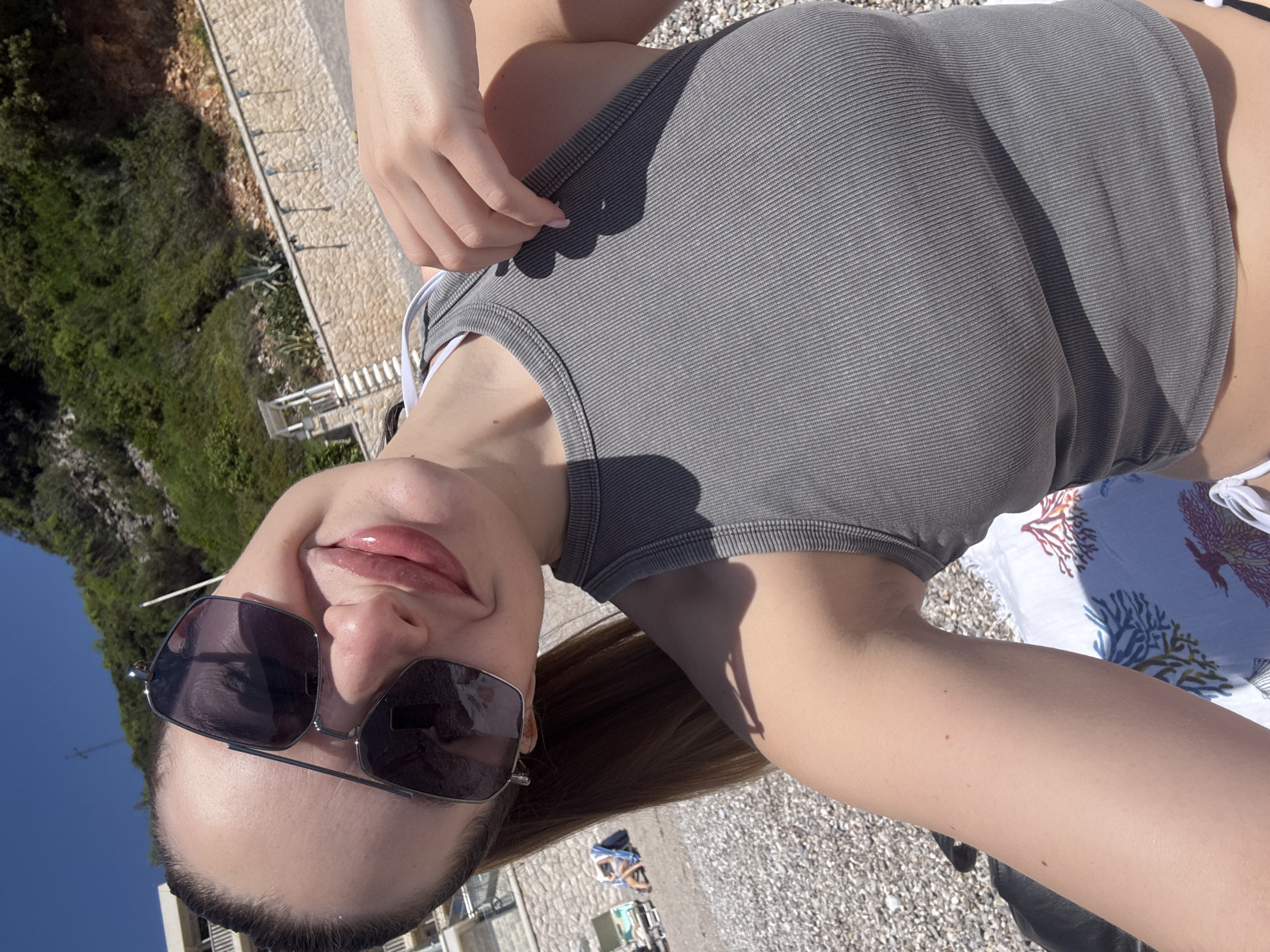 The image shows a woman on a beach in Prijevor, Montenegro. She is wearing sunglasses and a gray tank top. She is lying down on a towel. The beach is pebbly and there are some buildings in the background. There is vegetation on the hillside to the left. The sky is blue.