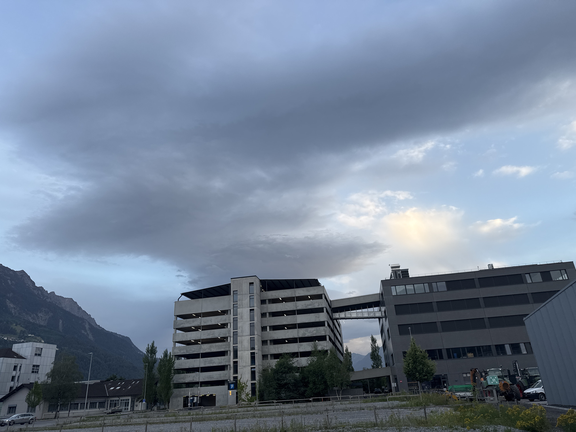 The image depicts a contemporary building complex situated in Nendeln, Liechtenstein, under a dynamic sky with a mix of dark, heavy clouds and lighter patches. The primary structures are a multi-level concrete parking garage and an adjacent, darker building, connected by an elevated enclosed walkway. The parking garage, with its open-air levels and exposed concrete structure, is the more prominent element on the left side of the frame. The darker building on the right features a modern facade with numerous dark-tinted windows.
To the left of the parking garage, a lower-rise building with white siding and dark-framed windows is visible, situated closer to the foreground. In the background, a large, densely forested mountain range dominates the horizon, its slopes partially obscured by atmospheric conditions. The sky above is a significant feature, with dramatic, textured clouds stretching across the upper portion of the image, suggesting an overcast or approaching weather system.
In the foreground, a gravelly, undeveloped area is present, with sparse vegetation and what appears to be a low fence or barrier. A silver-colored car is parked on the left side of the frame in front of the white building. On the right side of the frame, behind the darker building, several industrial vehicles, including a green tractor-like machine and what appears to be a smaller construction vehicle, are parked in an outdoor lot. The overall scene conveys an urban or industrial edge against a natural mountainous backdrop, under a moody sky.
