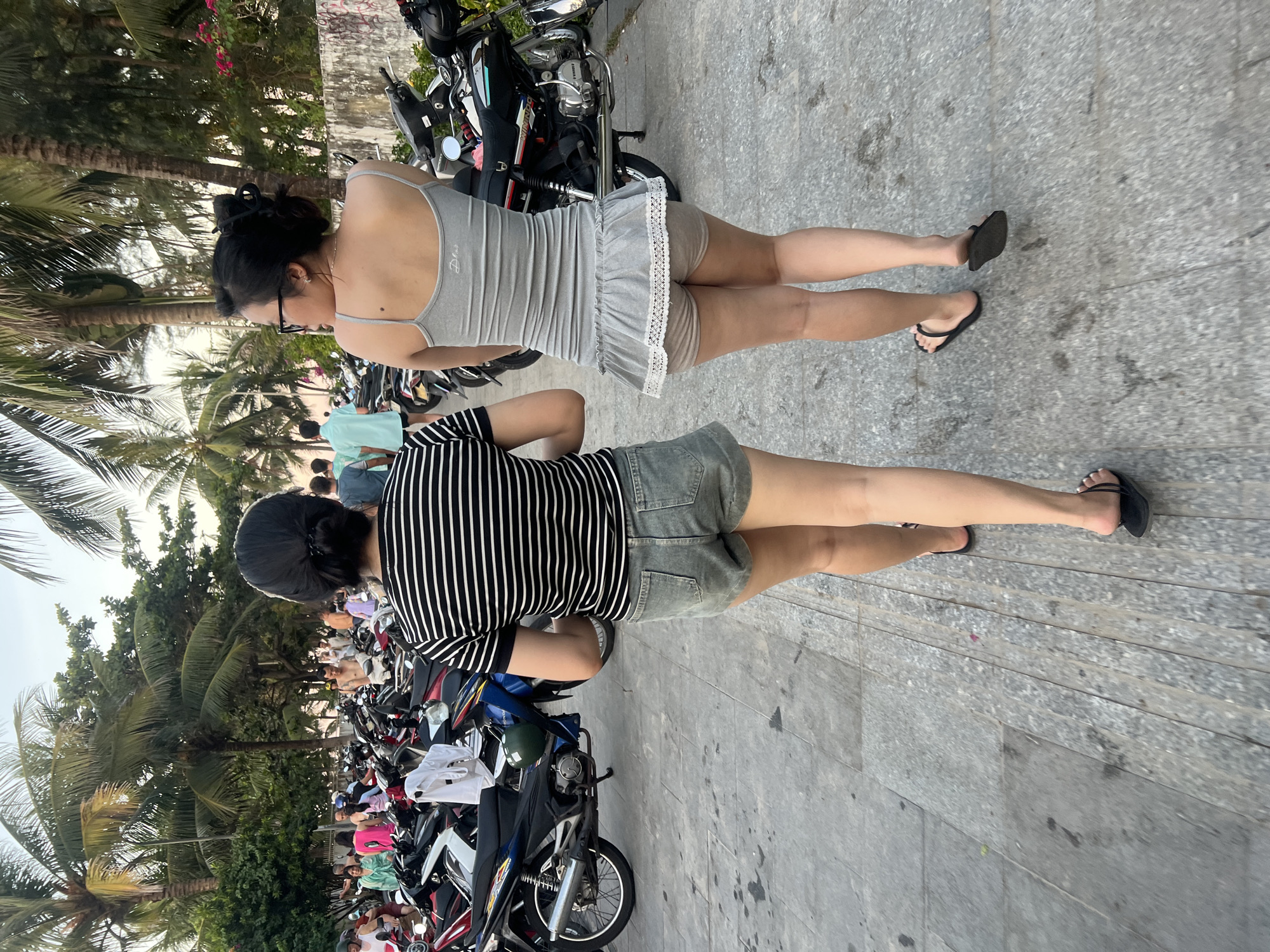 Two women walk away from the camera on a paved walkway lined with parked motorcycles. The woman on the left wears a black and white striped t-shirt and denim shorts. The woman on the right wears a grey tank top with a ruffled grey skirt and beige shorts. Both women wear black flip-flops. Palm trees and lush vegetation are visible in the background. The scene appears to be in a public area, possibly near a beach or a tourist attraction, given the presence of many motorcycles. The image was taken in Da Nang, Vietnam.