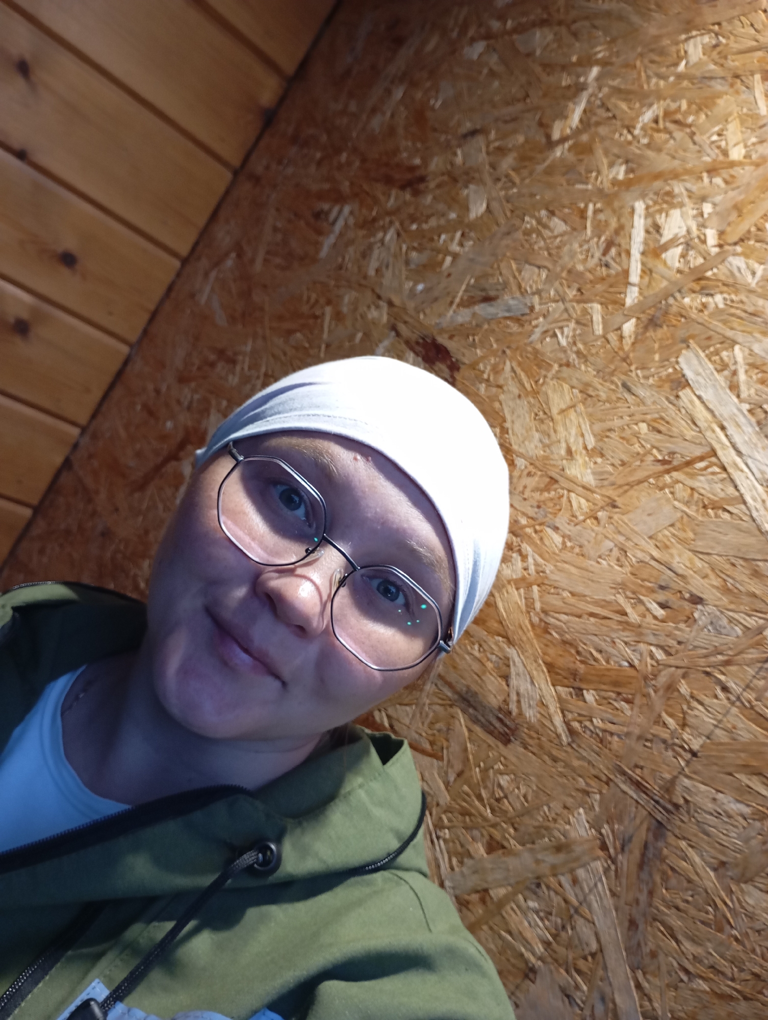 A medium shot captures a woman in a close-up selfie, her face angled towards the camera. She is wearing a white head covering and round glasses with thin, silver frames. Her eyes are blue and focused on the viewer. She has a faint smile on her lips. She is dressed in a green hooded jacket over a white t-shirt. The background consists of two distinct textures: on the left, a wall made of horizontal wooden planks, and on the right, a wall constructed from compressed wood chips, resembling OSB board. The lighting suggests an indoor environment, possibly a rustic dwelling in Malyi Bukor, Russia, given the materials used for the walls.