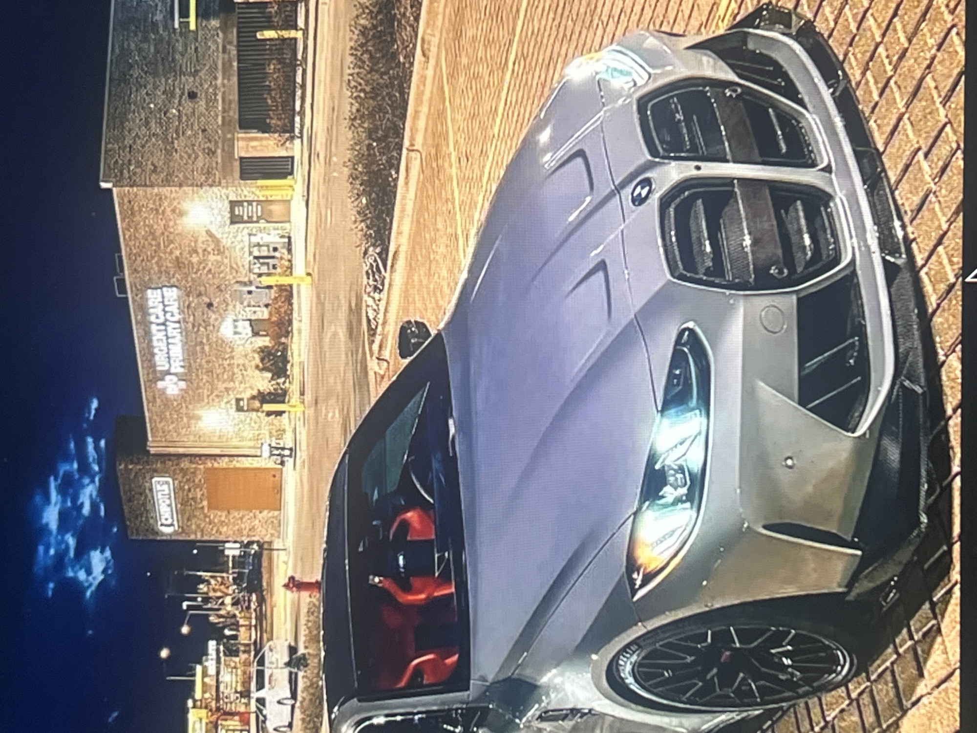In the city of Vanadzor, Armenia, at night, a silver BMW M4 is parked on a brick-paved surface in front of a commercial building. The building has a brick facade and features signage for "Chipotle" and "Urgent Care Primary Care." The car's interior has bright red racing seats visible through the windshield. The sky above is dark blue with some scattered clouds. The scene is lit by artificial lights from the building and streetlights, creating reflections on the car.