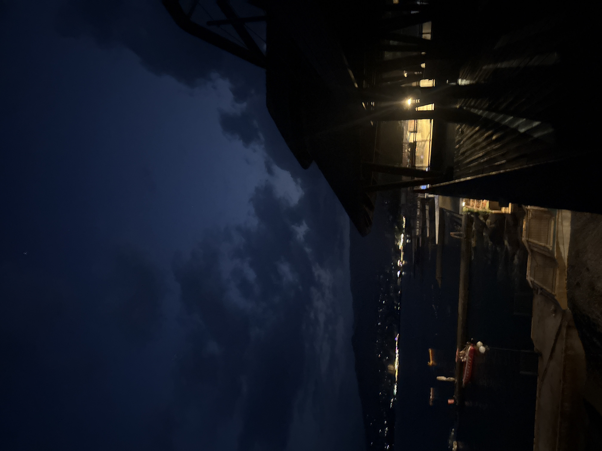 The image depicts a nighttime scene overlooking the city of Yalta, Ukraine, from a slightly elevated vantage point. The foreground is dominated by the dark silhouette of a roofed structure, possibly a gazebo or pavilion, with strong diagonal shadows cast by light sources within or behind it. The roof appears to be made of corrugated material. To the left of the structure, the dark water of the sea is visible, with several small boats moored or drifting. One red boat with white fenders is particularly noticeable. In the distance, the lights of Yalta twinkle along the coastline, with hills rising behind the city under a cloudy, dark blue sky. The overall atmosphere is moody and serene, characterized by low light conditions and the glow of distant city lights against the twilight sky.